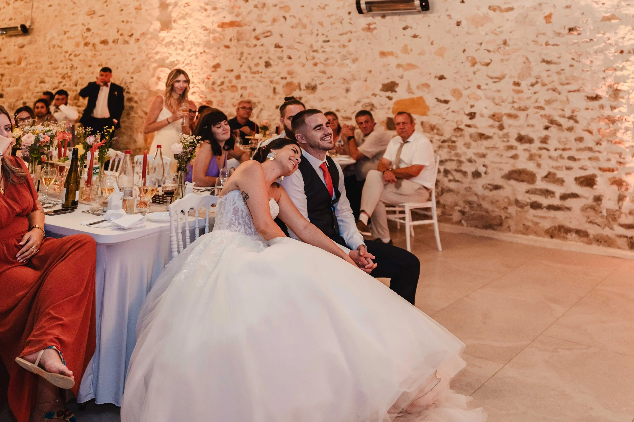 Un couple de mariés assis ensemble, la femme en robe de mariée blanche et le homme en costume, riant et partageant un moment heureux lors d'une réception de mariage dans une salle avec mur en pierre.
