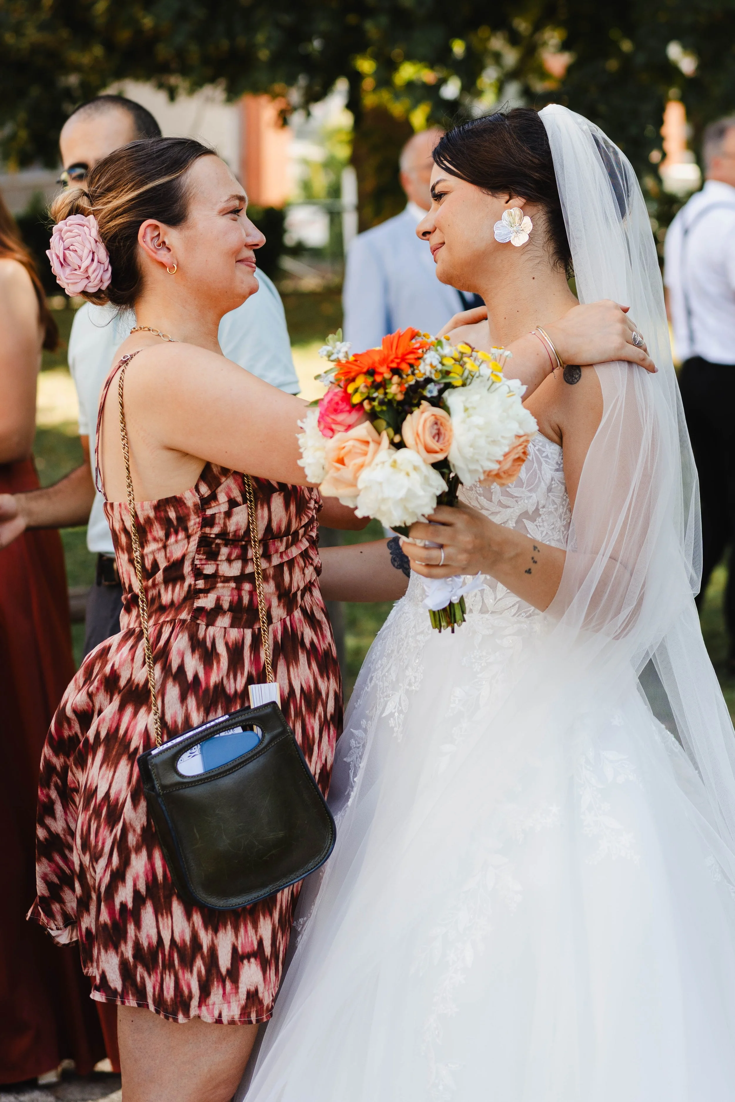 Deux femmes, dont une en robe de mariée, s'étreignent lors d'un mariage en extérieur, une d'elles tenant un bouquet de fleurs colorées.