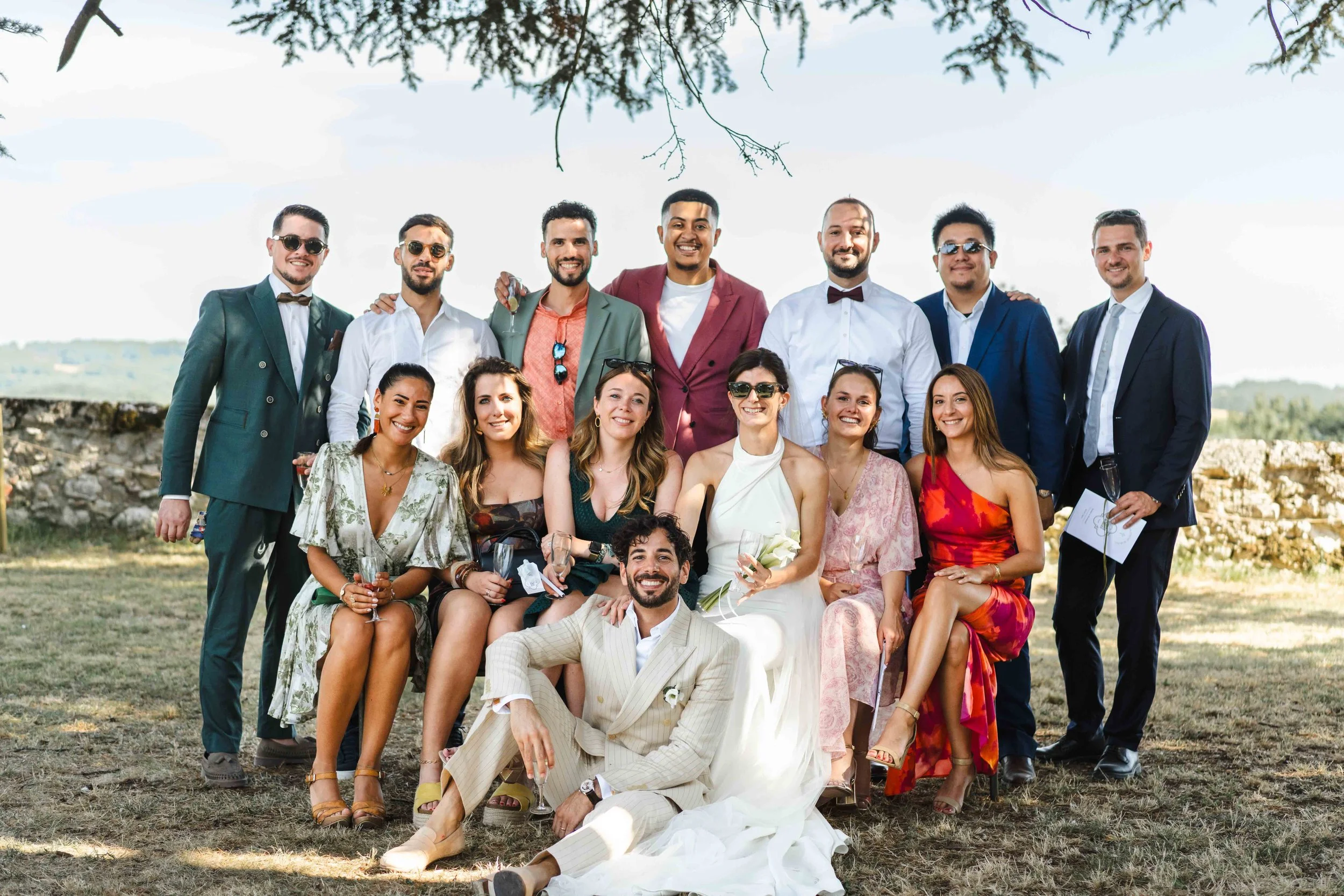 Groupe de personnes lors d'un mariage, posant en extérieur avec un ciel clair et un paysage champêtre en arrière-plan.