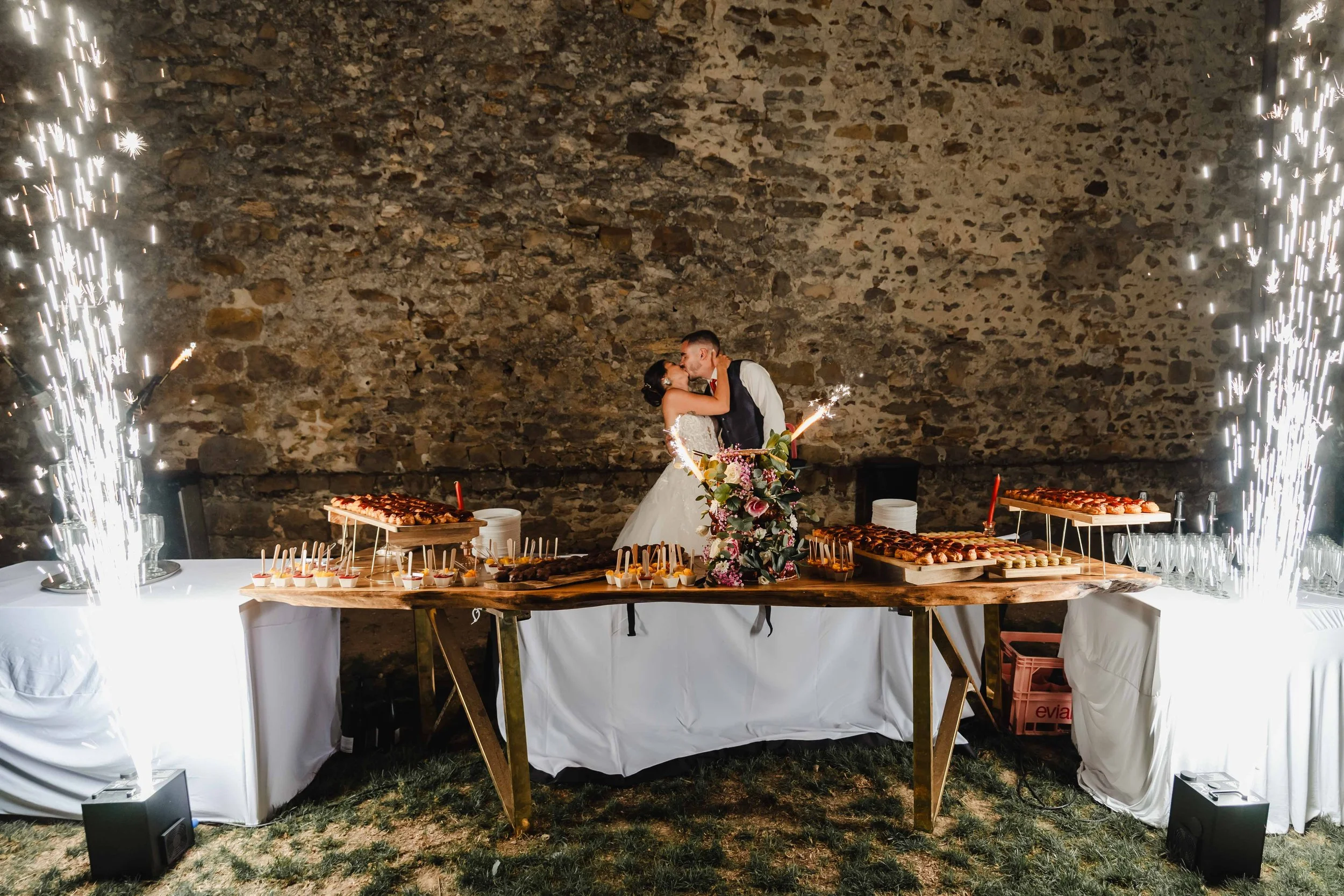 Un couple de mariés s'embrasse devant un buffet de fêtes avec des feux d'artifice, dans un décor rustique en pierre.