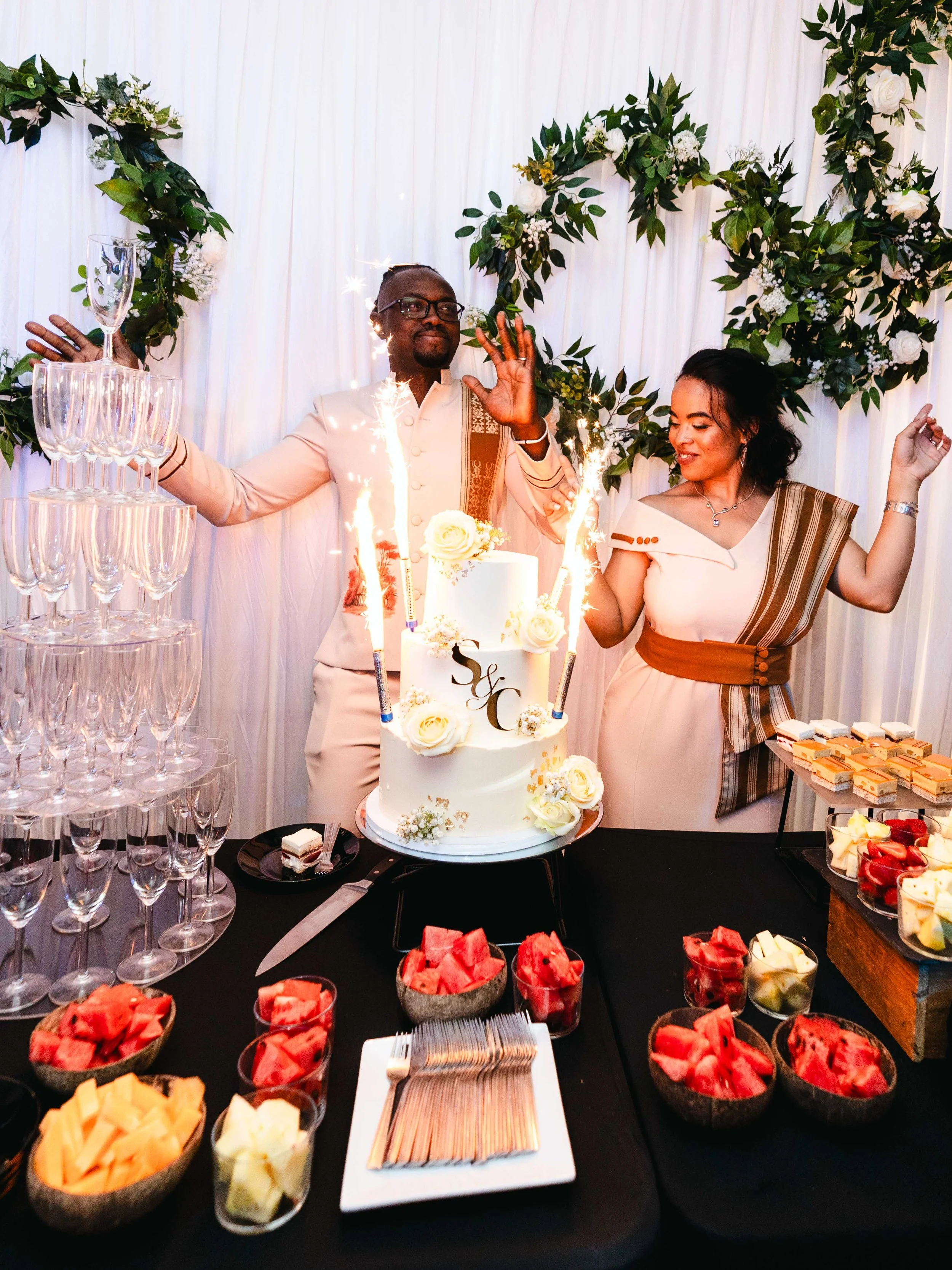 Un couple célèbre leur mariage avec un gâteau blanc décoré de fleurs, des feux d'artifice et une pièce montée de coupes à champagne vides, entourés d'une décoration florale.