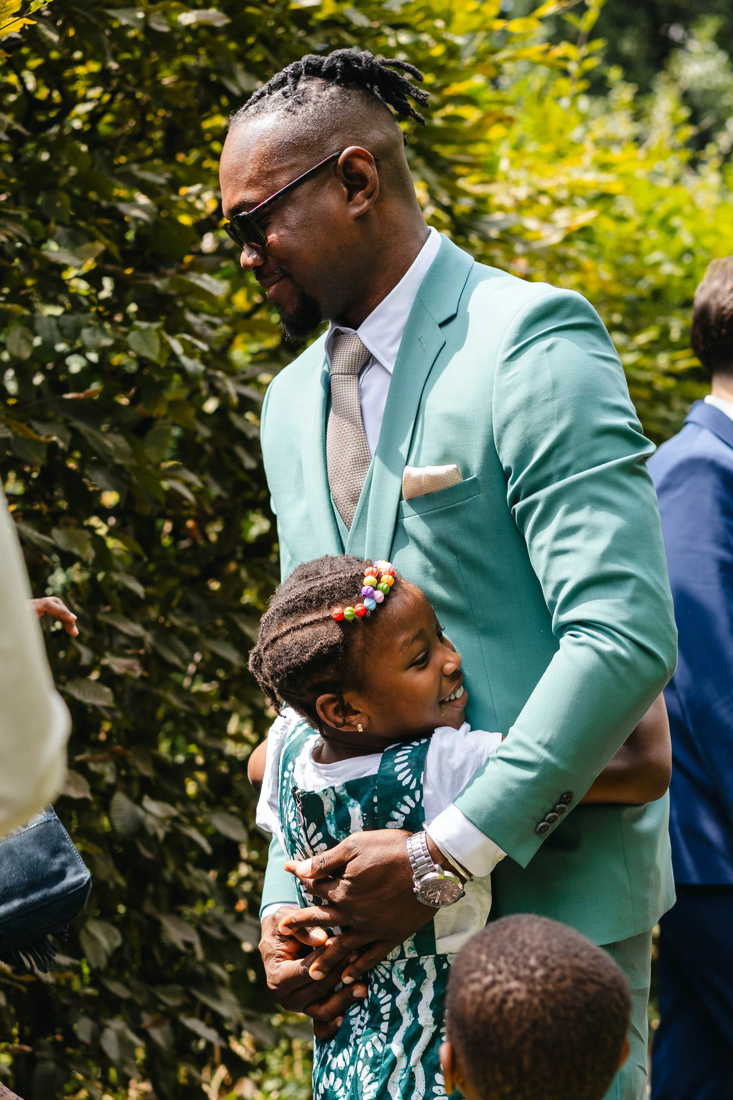 Un homme en costume vert clair embrasse une jeune fille lors d’un événement extérieur, avec des personnes en arrière-plan.