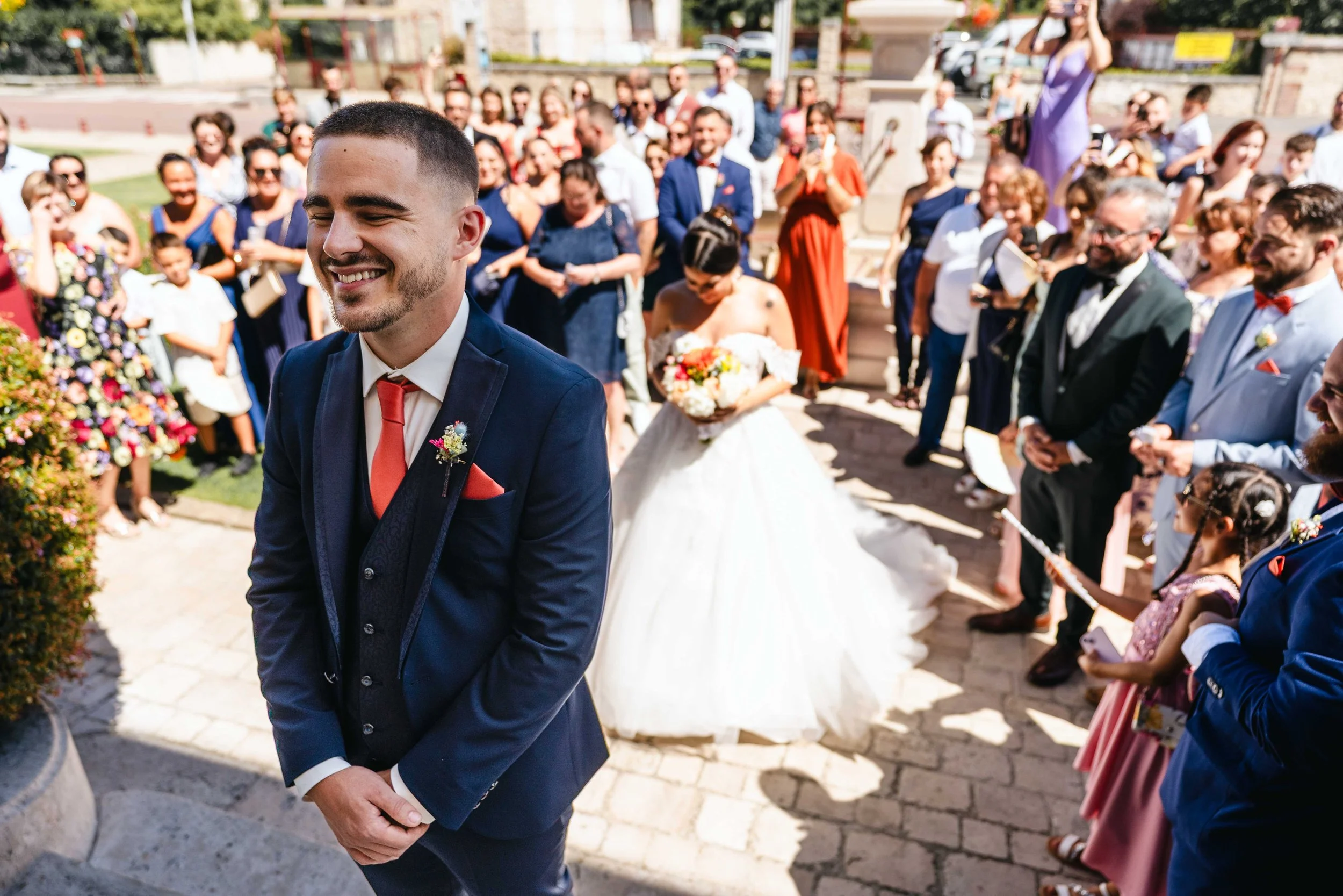 Un marié souriant en costume bleu avec une cravate rouge, devant un cortège de mariage, femmes et enfants en tenues colorées, lors d'une cérémonie en plein air ensoleillée à Bagneux Sur Loing.