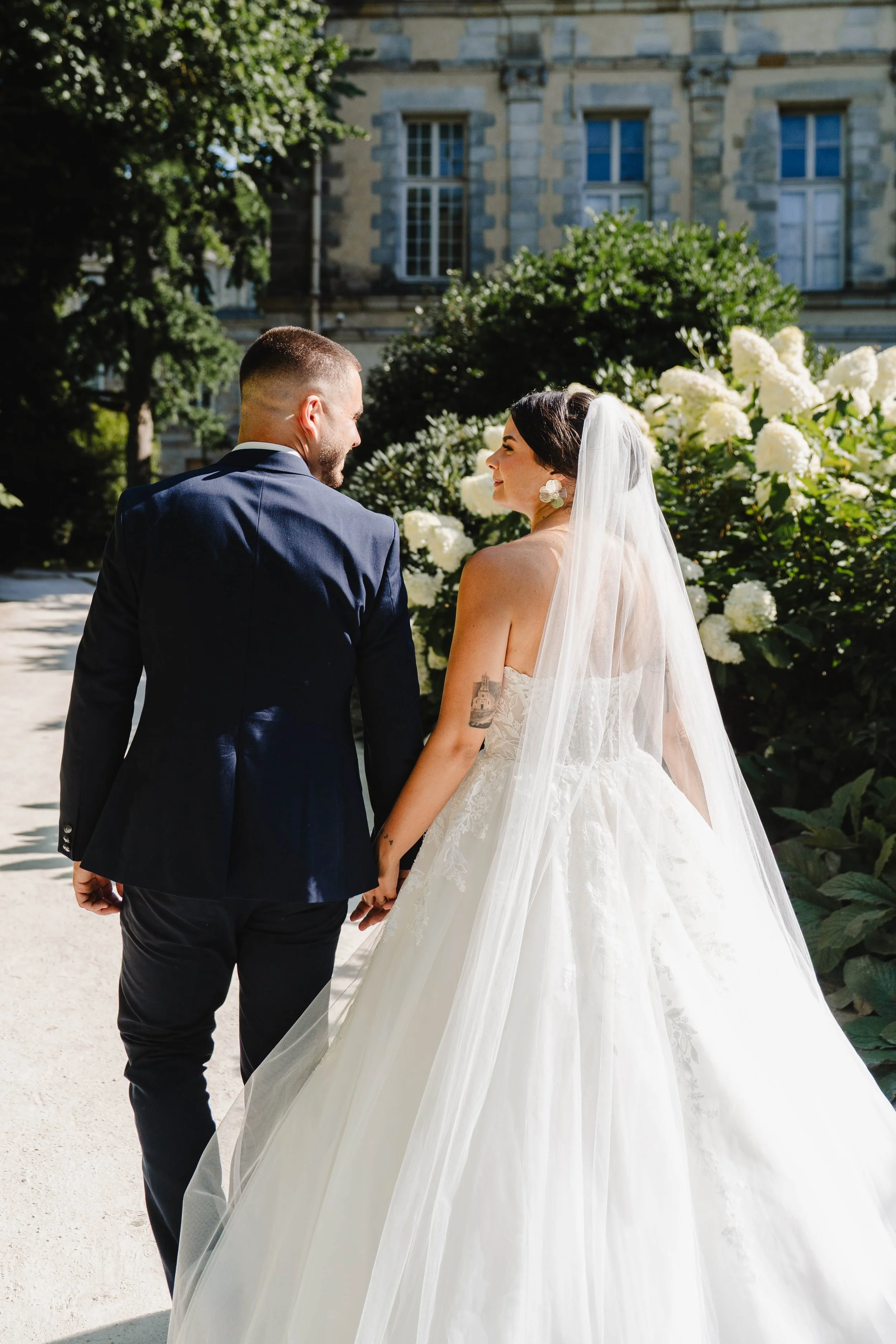 Un couple en mariage marche main dans la main dans un jardin fleuri, vu de l'arrière, avec une vieille bâtisse en pierre en fond, ensoleillé.