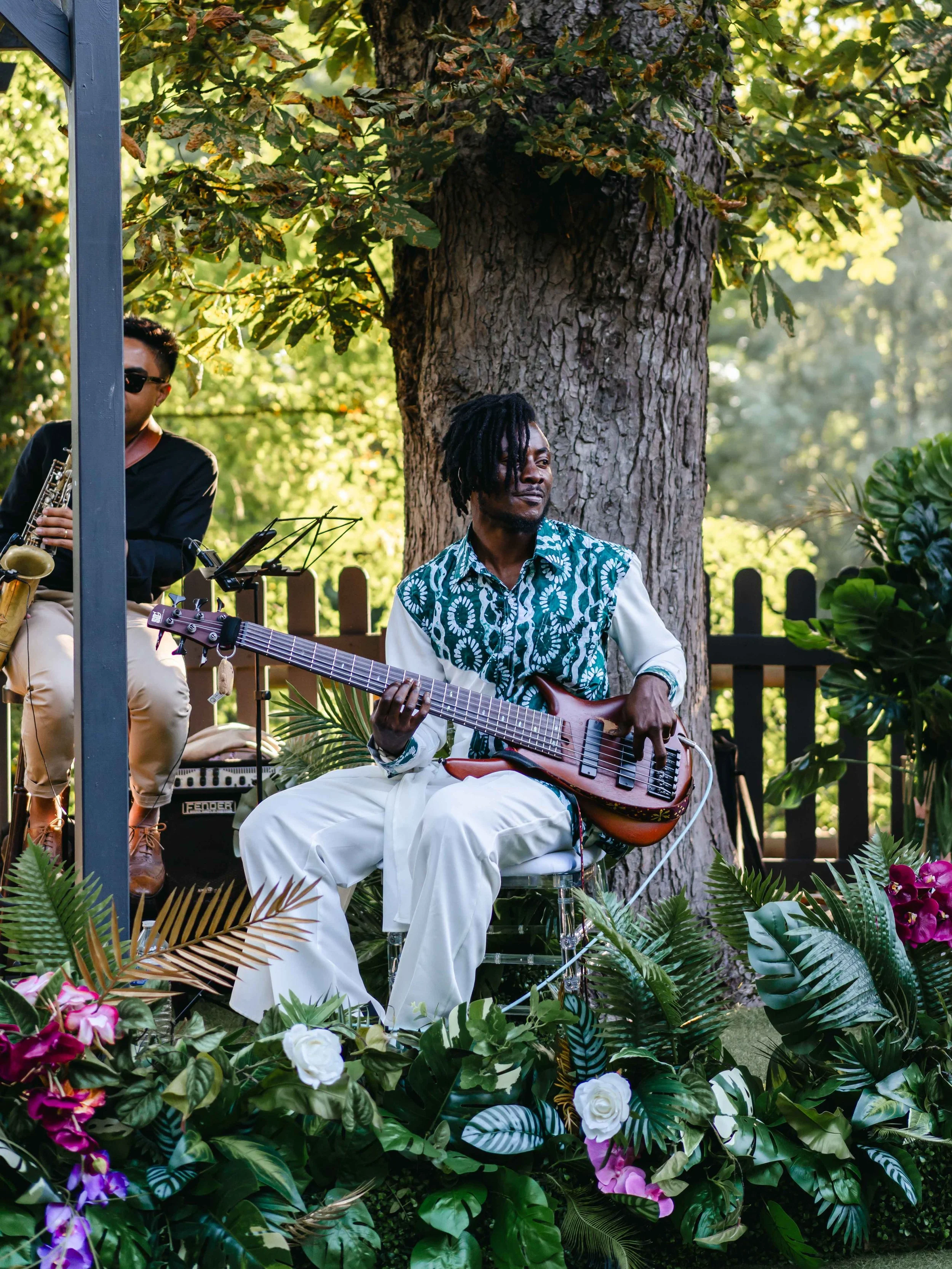 Musicien jouant de la guitare électrique lors d'une performance en plein air, entouré de plantes et d'arbres, avec un autre musicien jouant du saxophone à proximité.