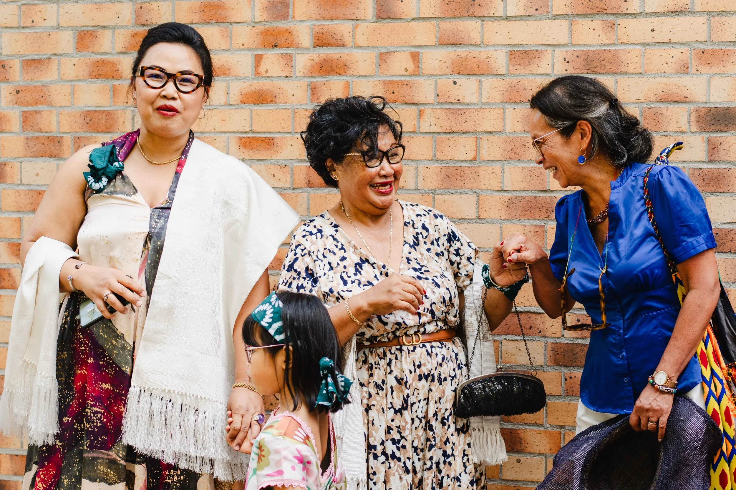 Trois femmes et une enfant, riant et se saluant, devant un mur en briques, lors d'une rencontre amicale.
