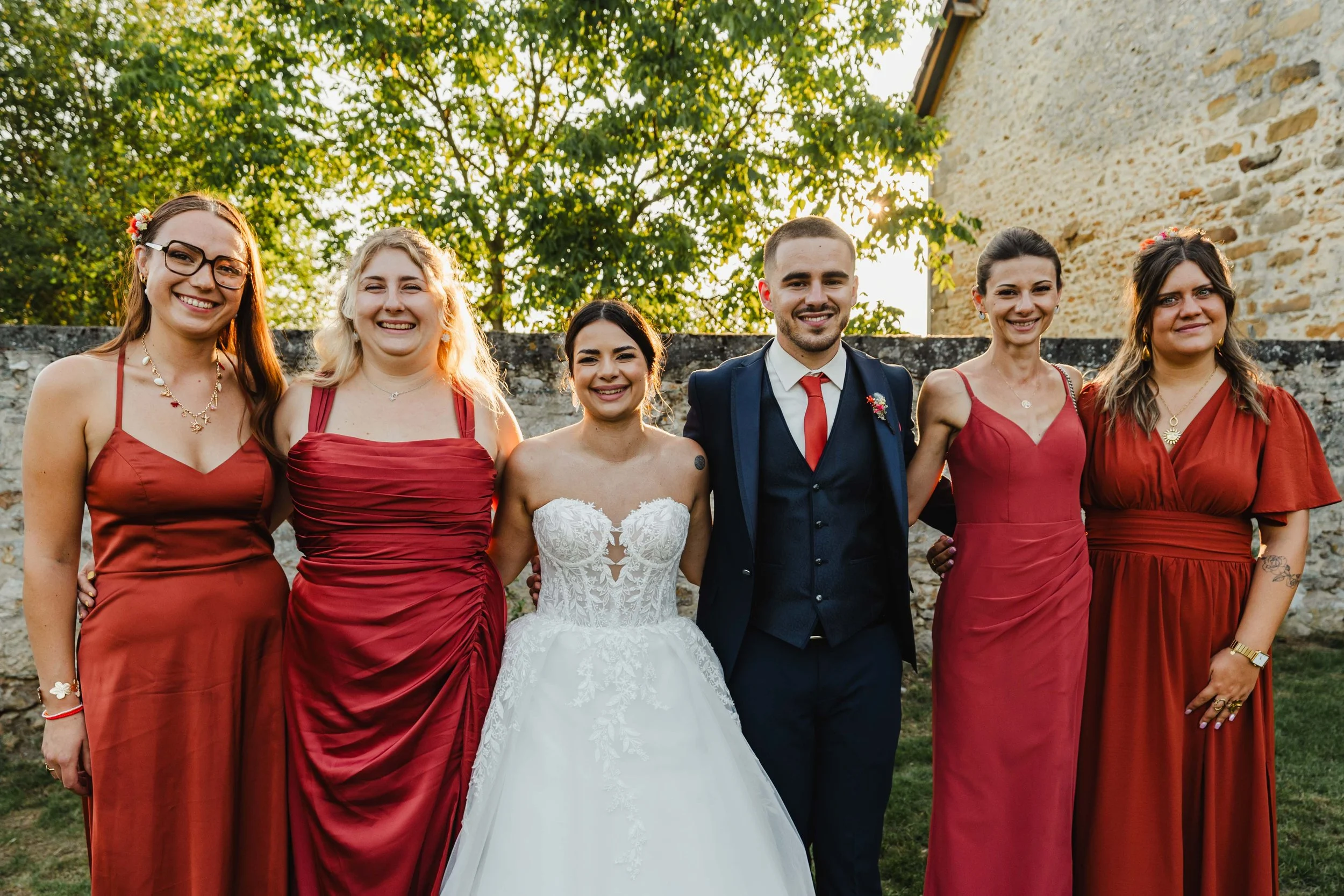 Groupe de six personnes en robes de mariée et costumes lors d'un mariage en plein air, sous un arbre, en fin d'après-midi.