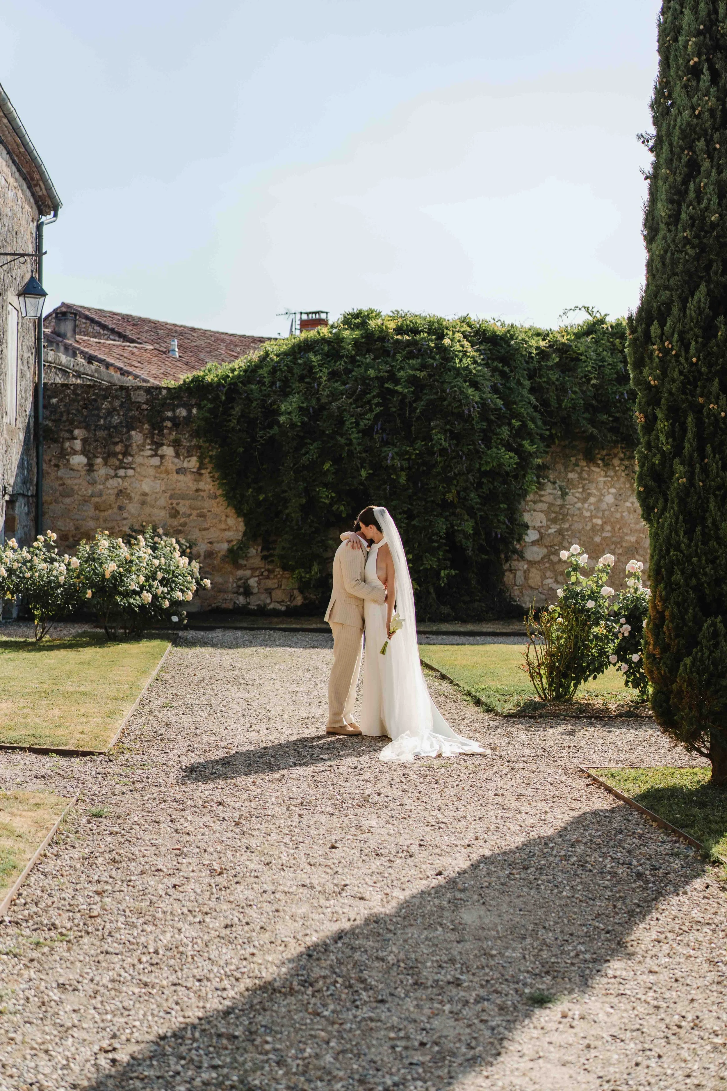 Un couple en mariage se tient dans un jardin, se regardant tendrement, avec un mur en pierre et des arbustes en fleurs derrière eux.