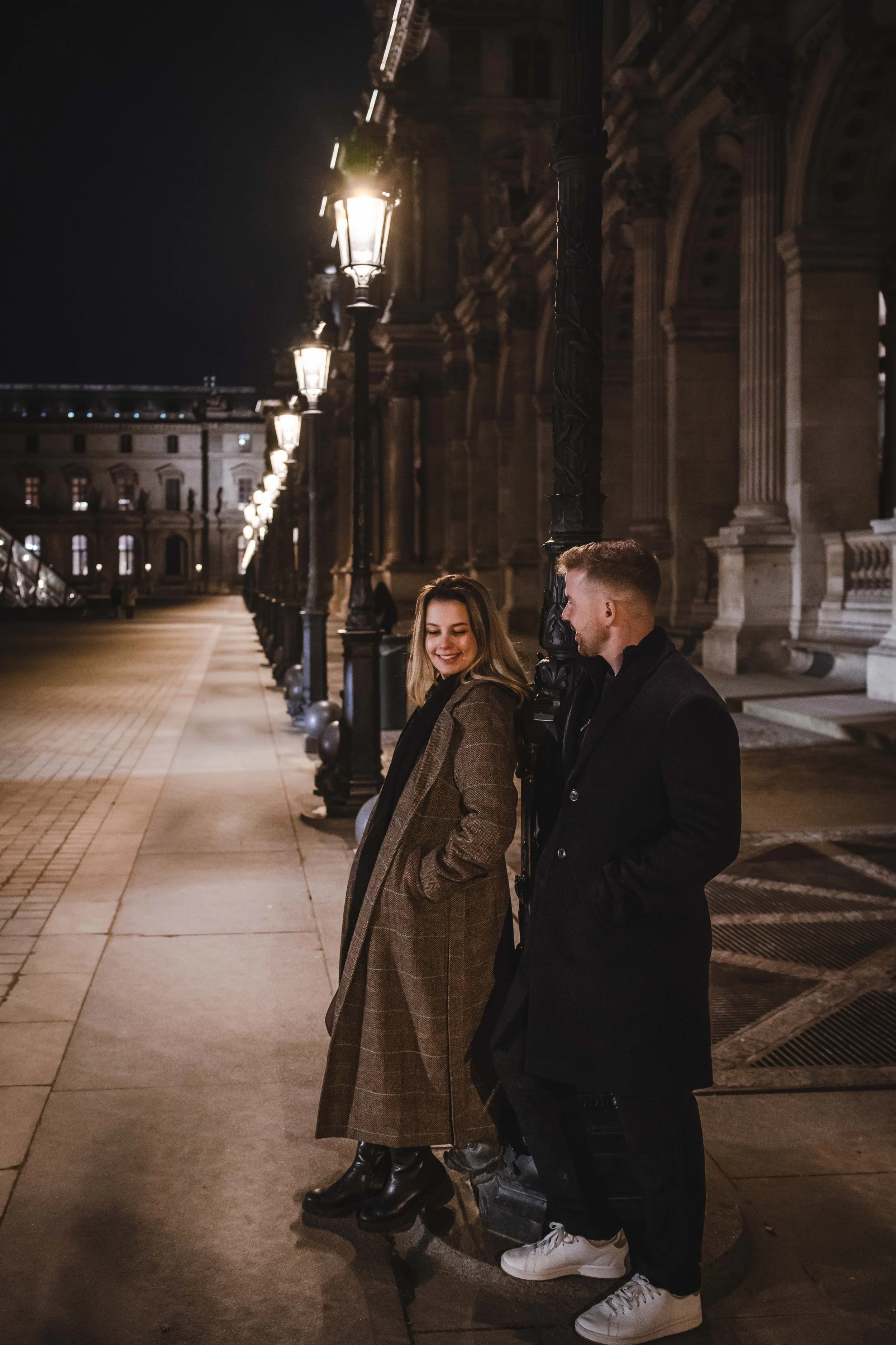 Un homme et une femme discutent et rient dans une rue éclairée la nuit, en face d'un bâtiment historique, le Louvre avec des colonnes et des lampadaires.