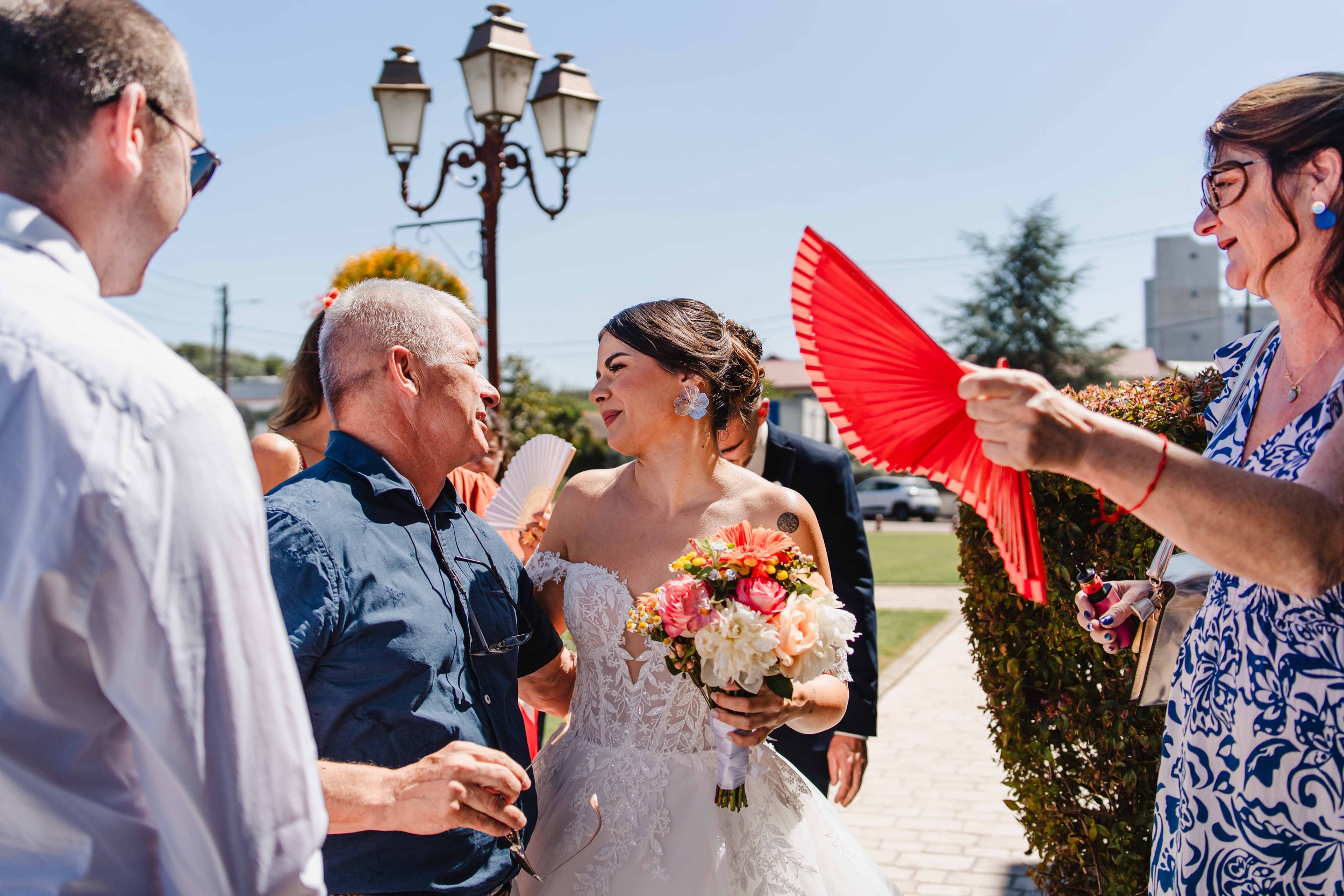Une mariée en robe blanche sourit face à un homme plus âgé lors d'une célébration en plein air, entourée de personnes tenant des éventails rouges et blancs, sous un ciel ensoleillé.