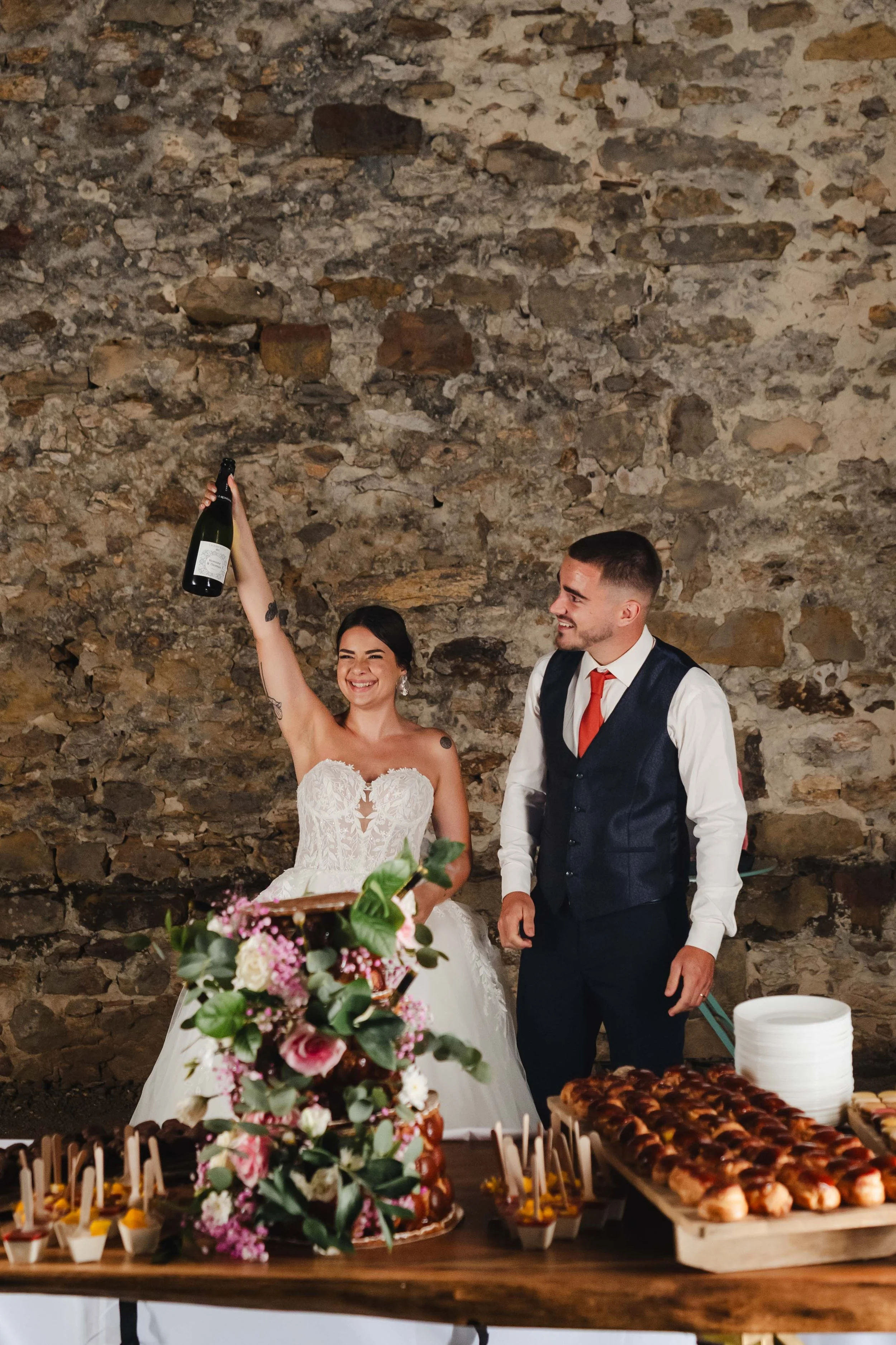 Un couple de mariés souriant à une fête, la mariée tenant une bouteille de champagne en l'air, devant une table décorée avec un gâteau de mariage, des fleurs et des desserts, dans une ambiance chaleureuse.