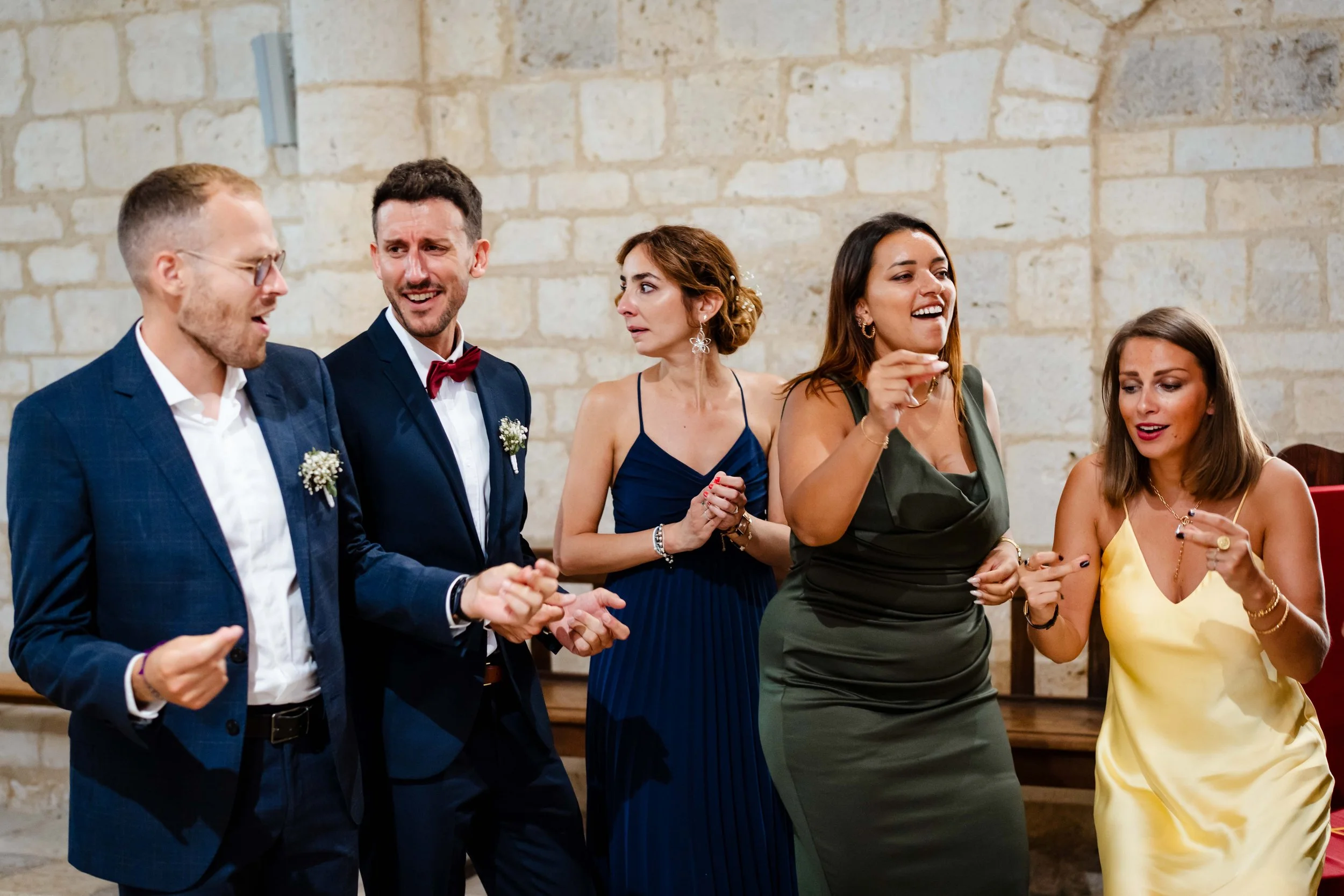 Cinq personnes habillées élégamment lors d'une fête ou d'un événement, semblant danser ou discuter, devant un mur en pierre.