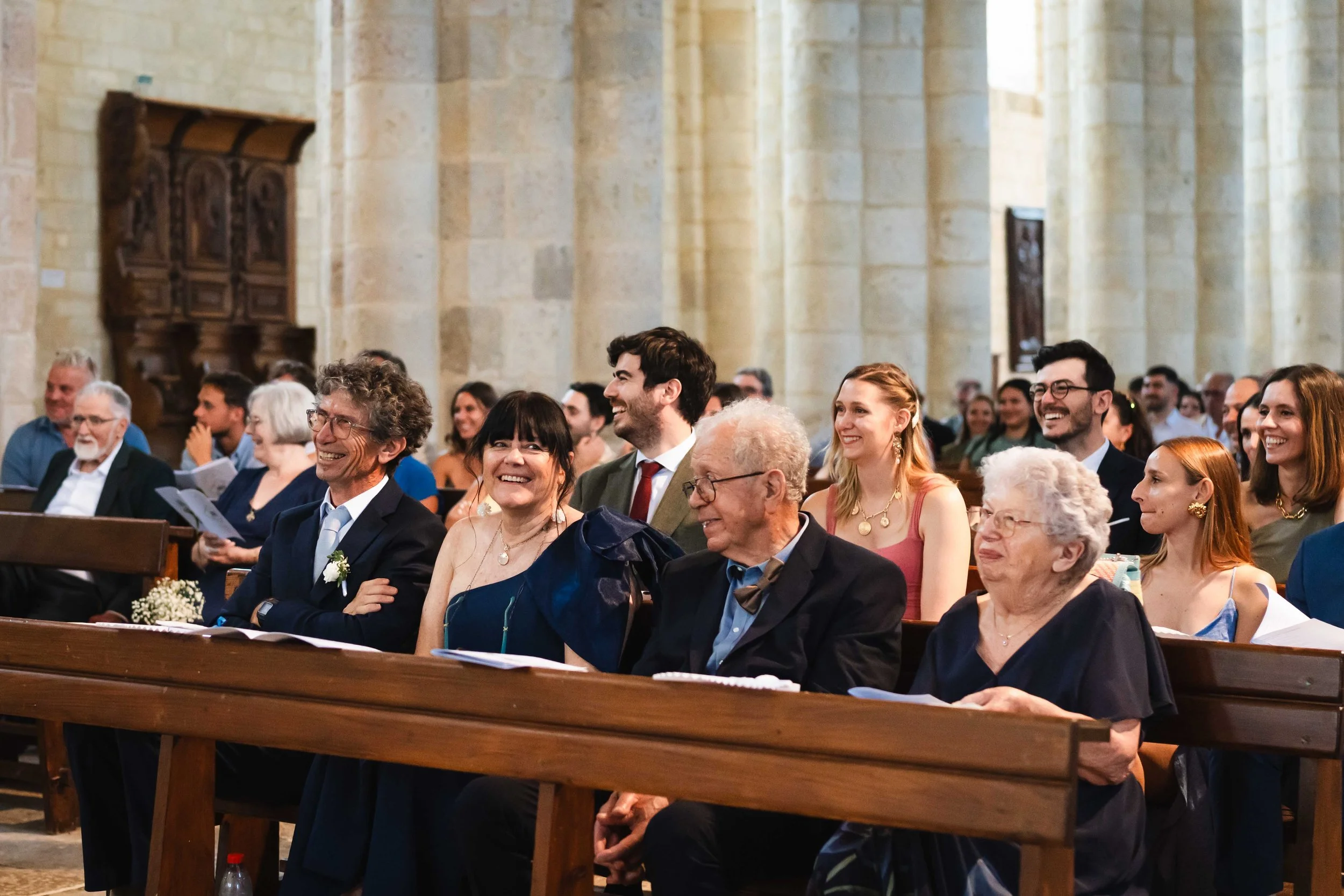 Gens assis dans une église, souriant et riant lors d'une cérémonie, probablement un mariage, dans un intérieur en pierre avec des bancs en bois.