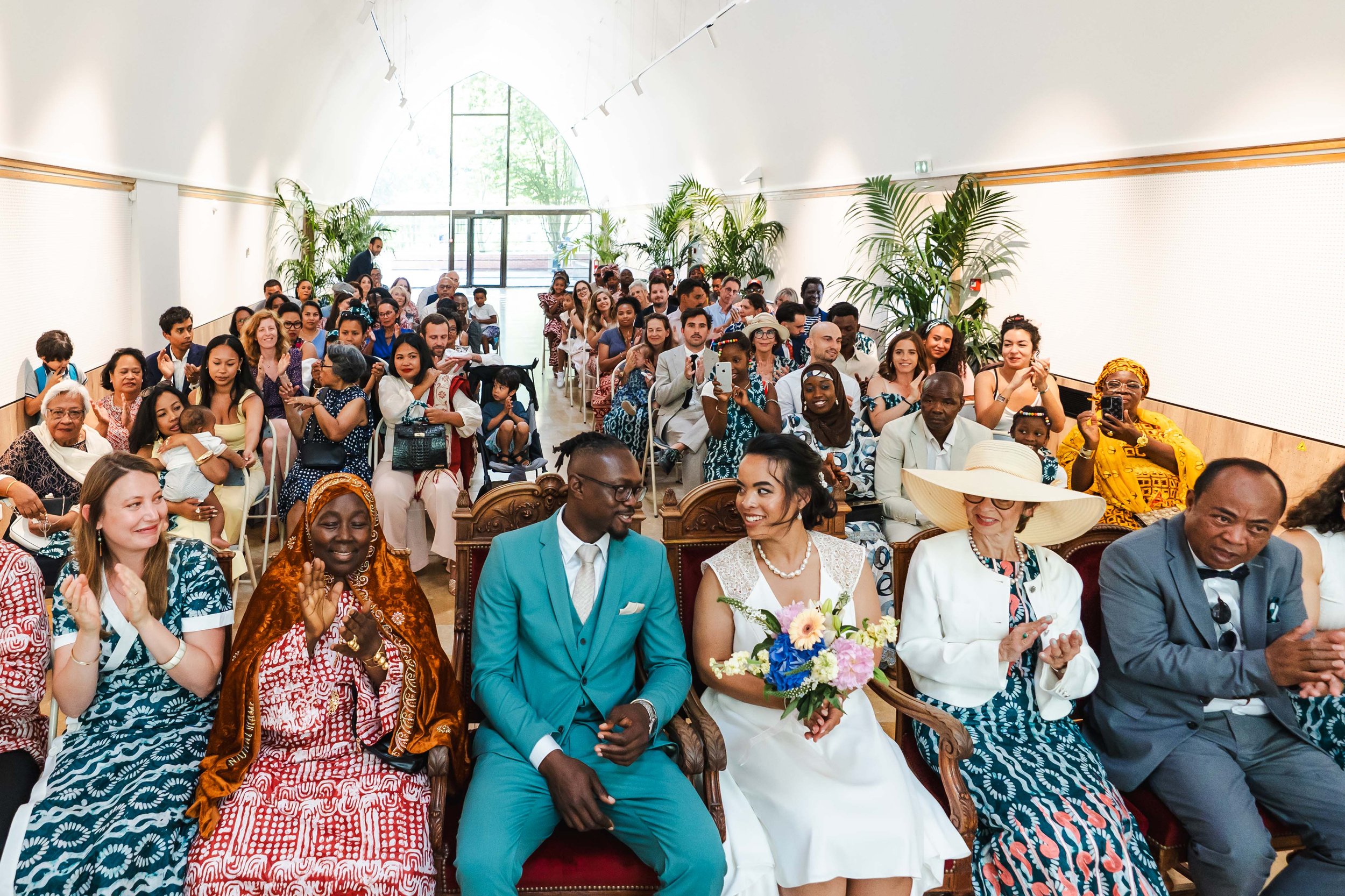 Vue de la cérémonie de mariage avec un couple assis devant le public, entouré de personnes diverses applaudissant et prenant des photos dans un lieu lumineux avec des plantes verticales.
