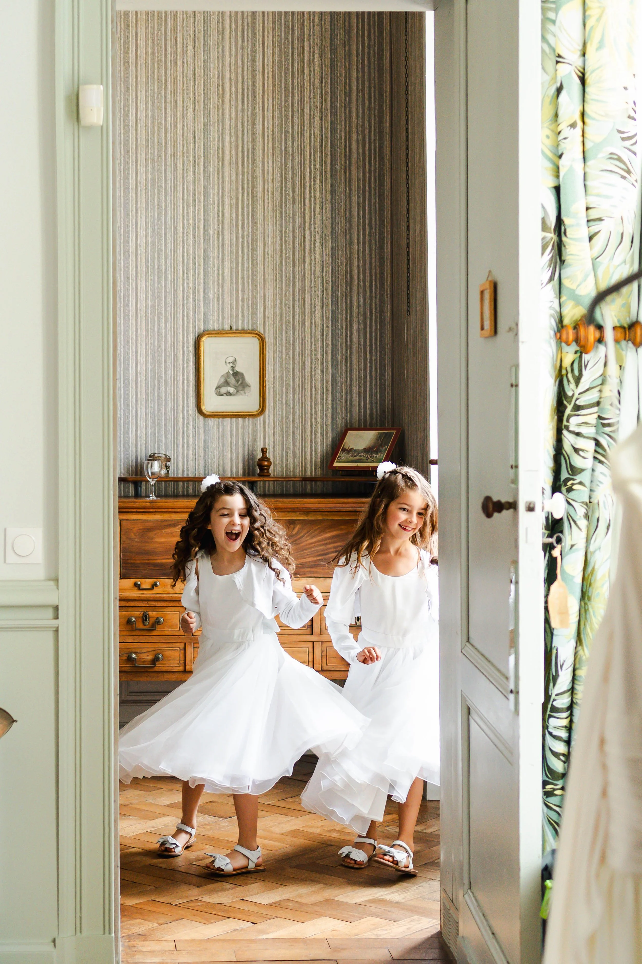 Deux jeunes filles habillées en robes blanches, souriantes et joyeuses, dans une pièce avec un meuble en bois et des photos encadrées, en train de jouer ou de danser.