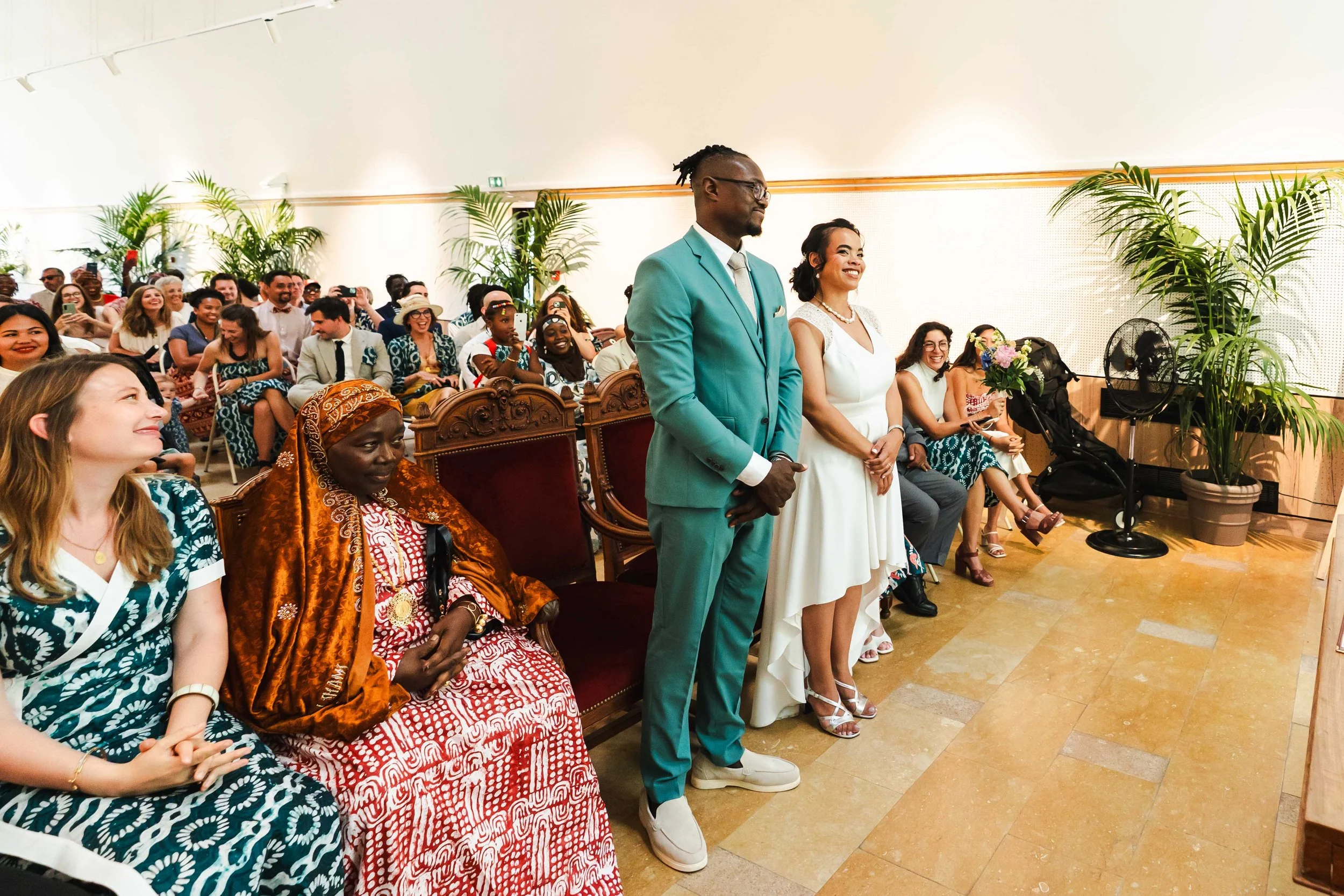Un couple en tenue de mariage se tient debout devant une assemblée lors d'une cérémonie de mariage. Autour d'eux, des invités s'assoient et prennent des photos, certains sourient et certains rient. La scène se déroule dans une salle décorée avec des 