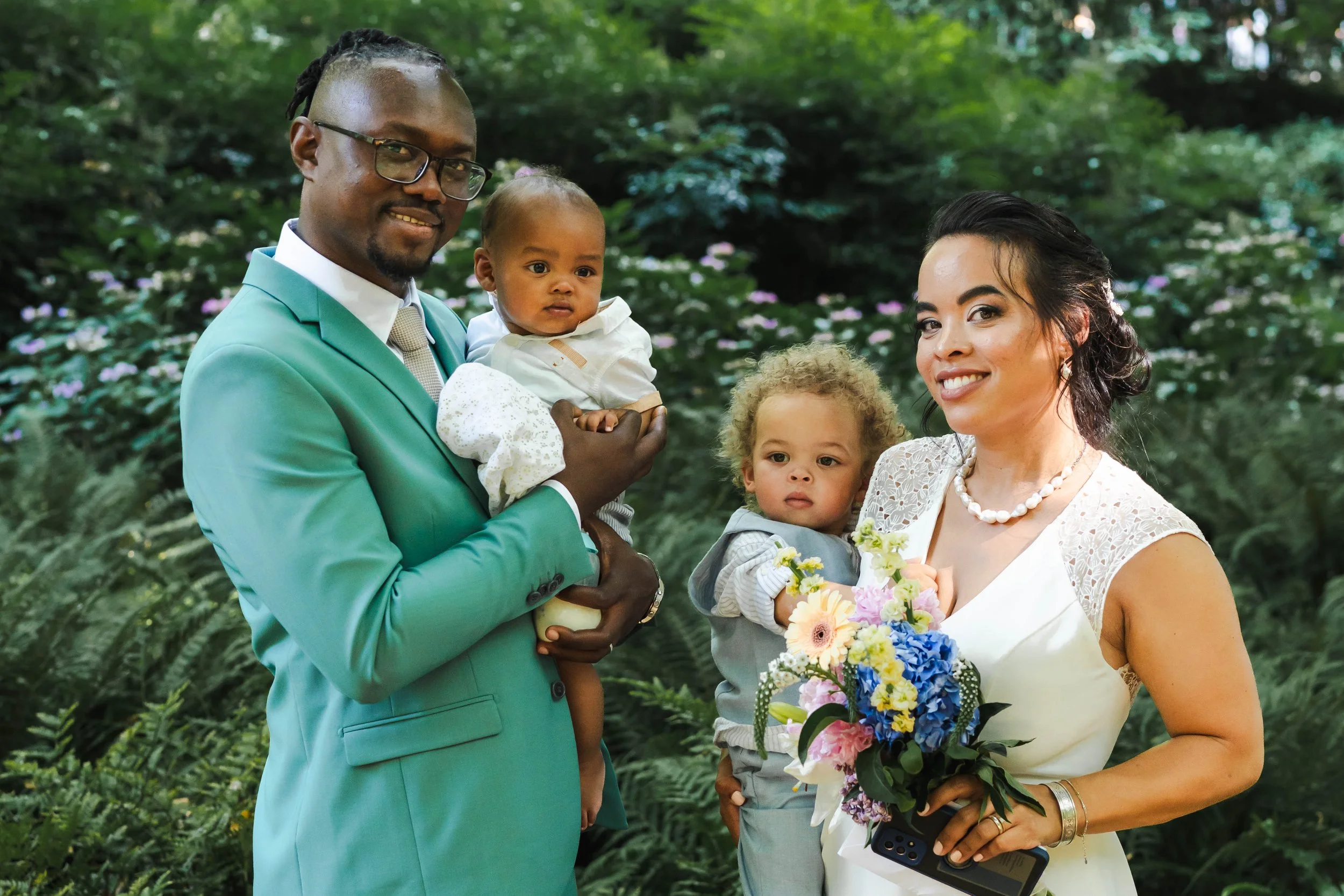 Un couple avec deux enfants dans un parc, la femme tient un bouquet de fleurs. L'homme porte un costume vert, et la femme porte une robe blanche avec un collier.