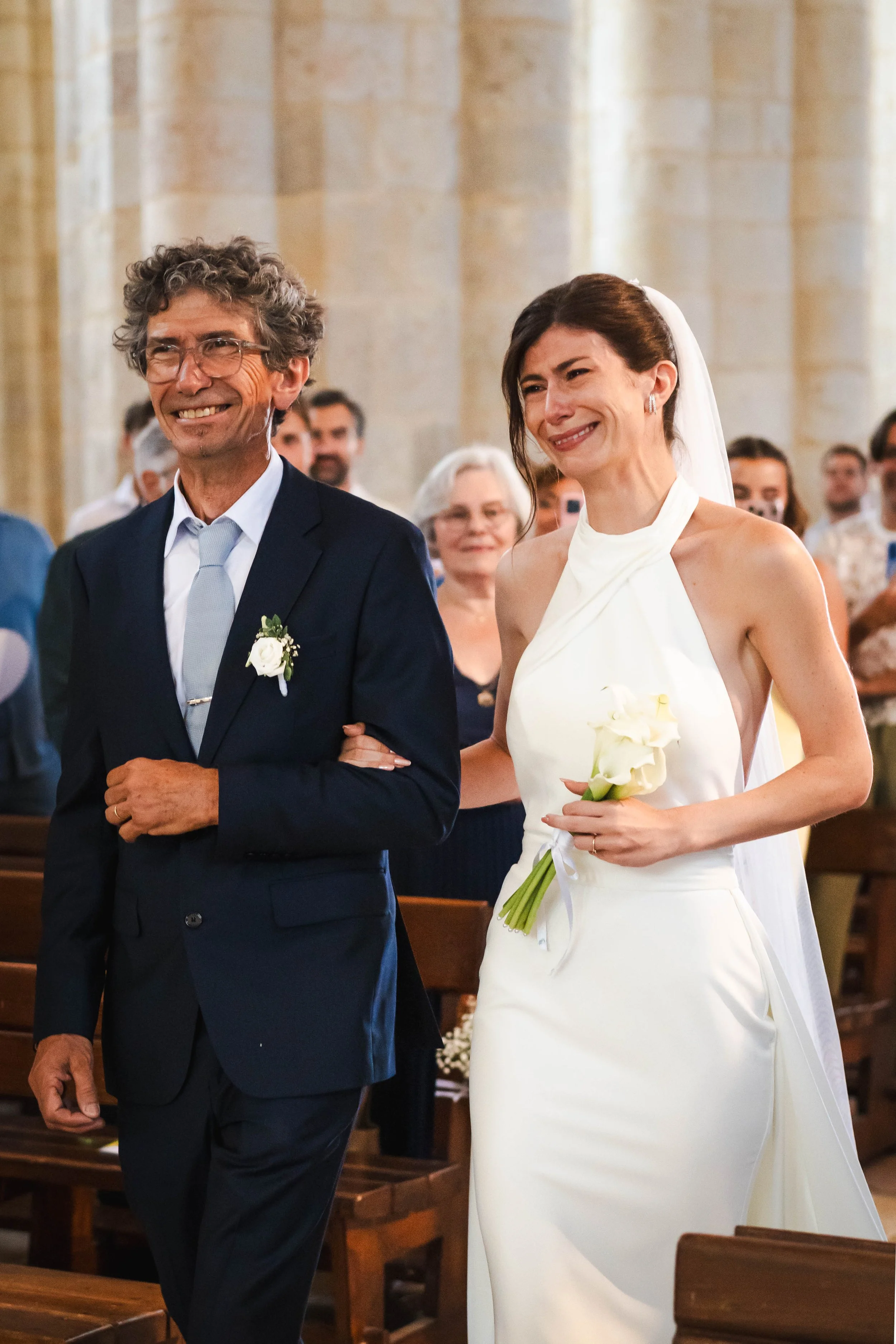 Une femme en robe blanche de mariage marche dans une église, soutenue par un homme en costume sombre, entourés de convives regardant la cérémonie.