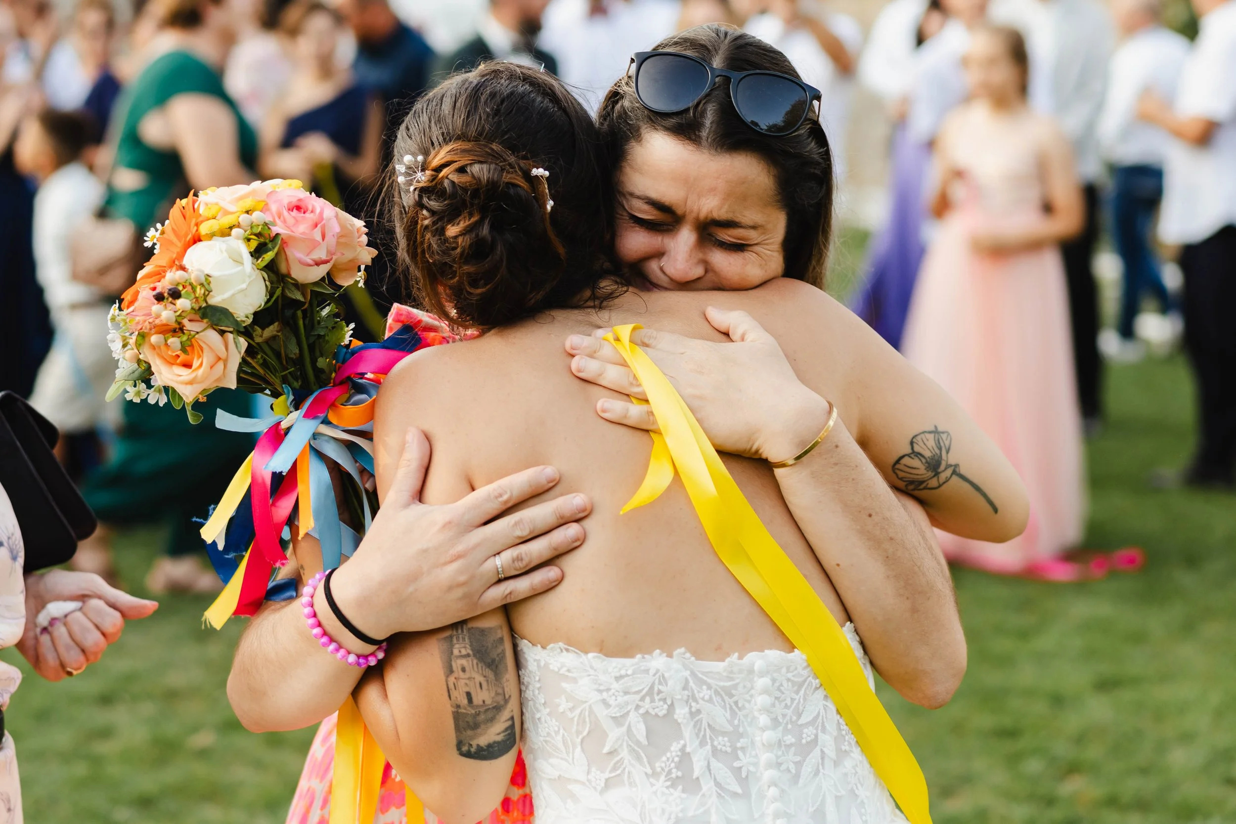 Deux femmes s'étreignent avec émotion lors d'un événement en plein air, une portant un bouquet de fleurs, l'autre en robe blanche avec des tatouages.