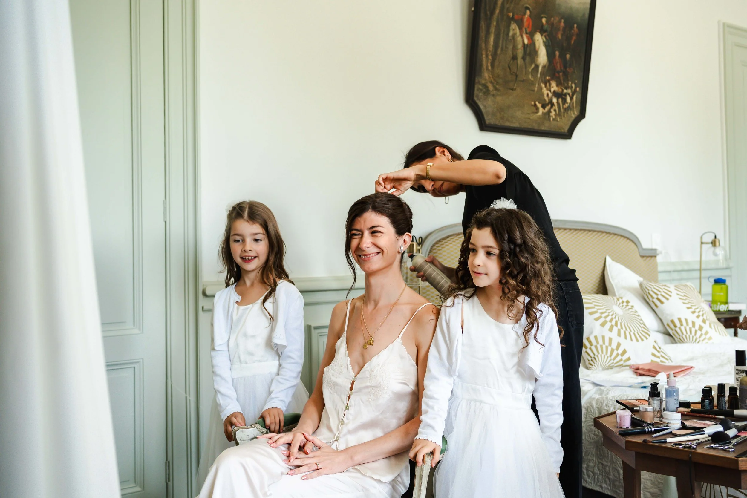 Une femme et deux jeunes filles en robe blanche sont en train de se faire maquiller et coiffer dans une chambre élégante, probablement en préparation pour un événement spécial.