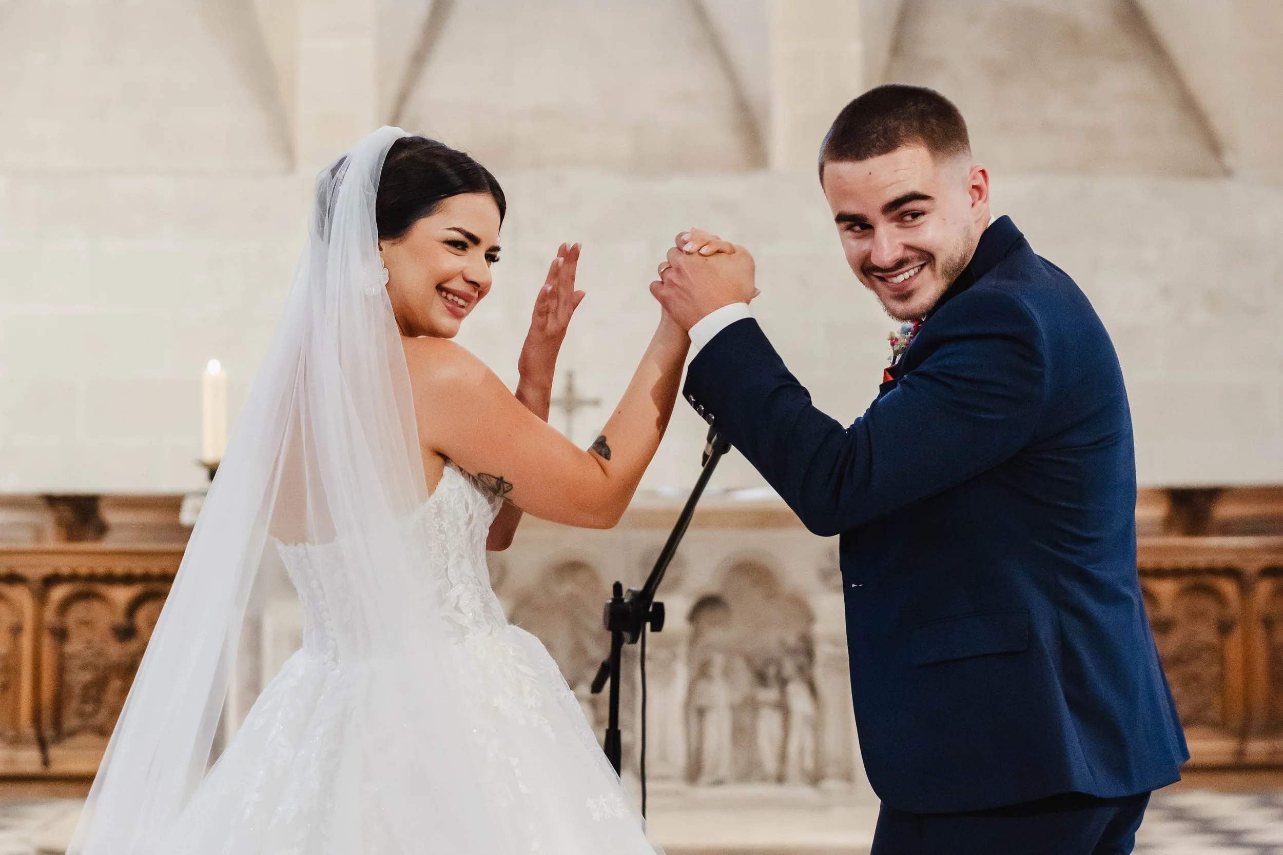 Un couple de mariés danse lors de leur mariage dans une église, la bride porte une robe blanche avec plume et le marié porte un costume bleu foncé. Ils sourient et s'amusent, la bride porte une voile blanche et des boucles d'oreilles. L'arrière-plan 