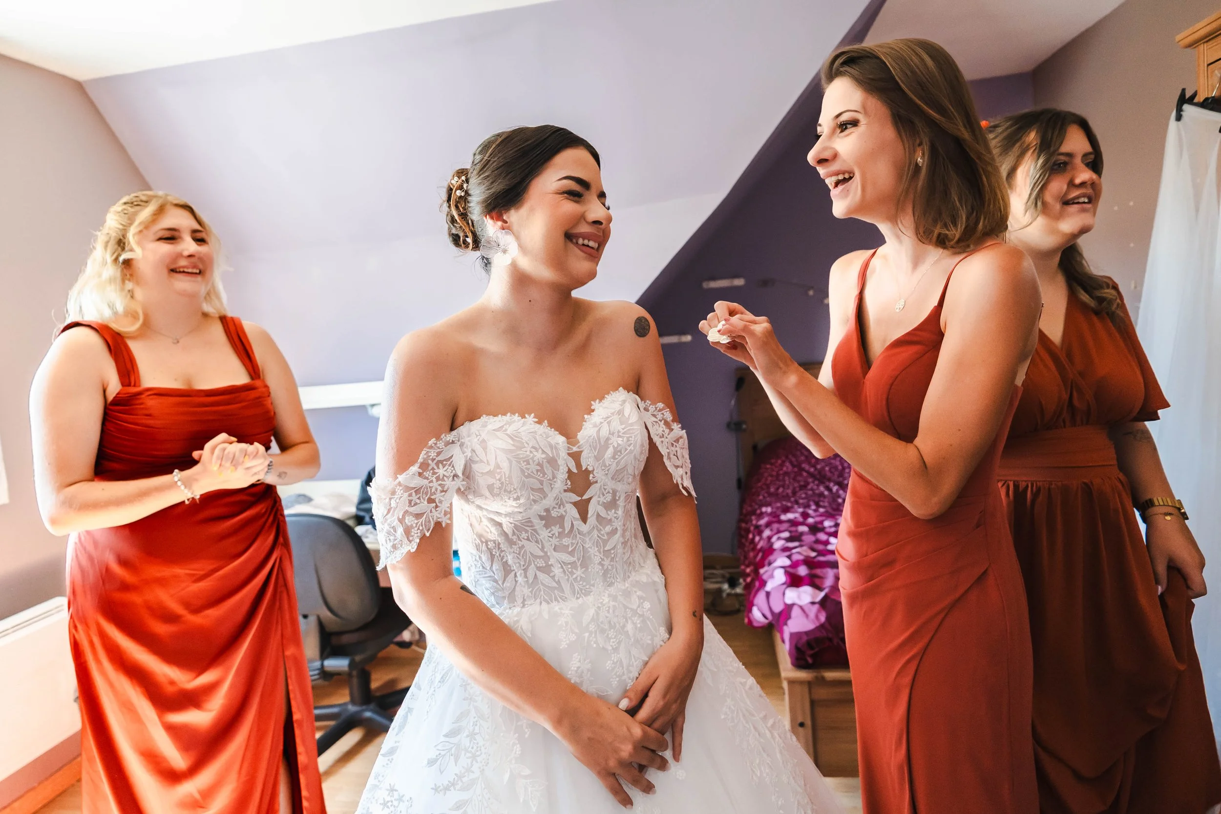 Une femme en robe de mariée souriant et parlant avec une amie en robe rouge, entourées d'autres femmes en robes rouges dans une pièce