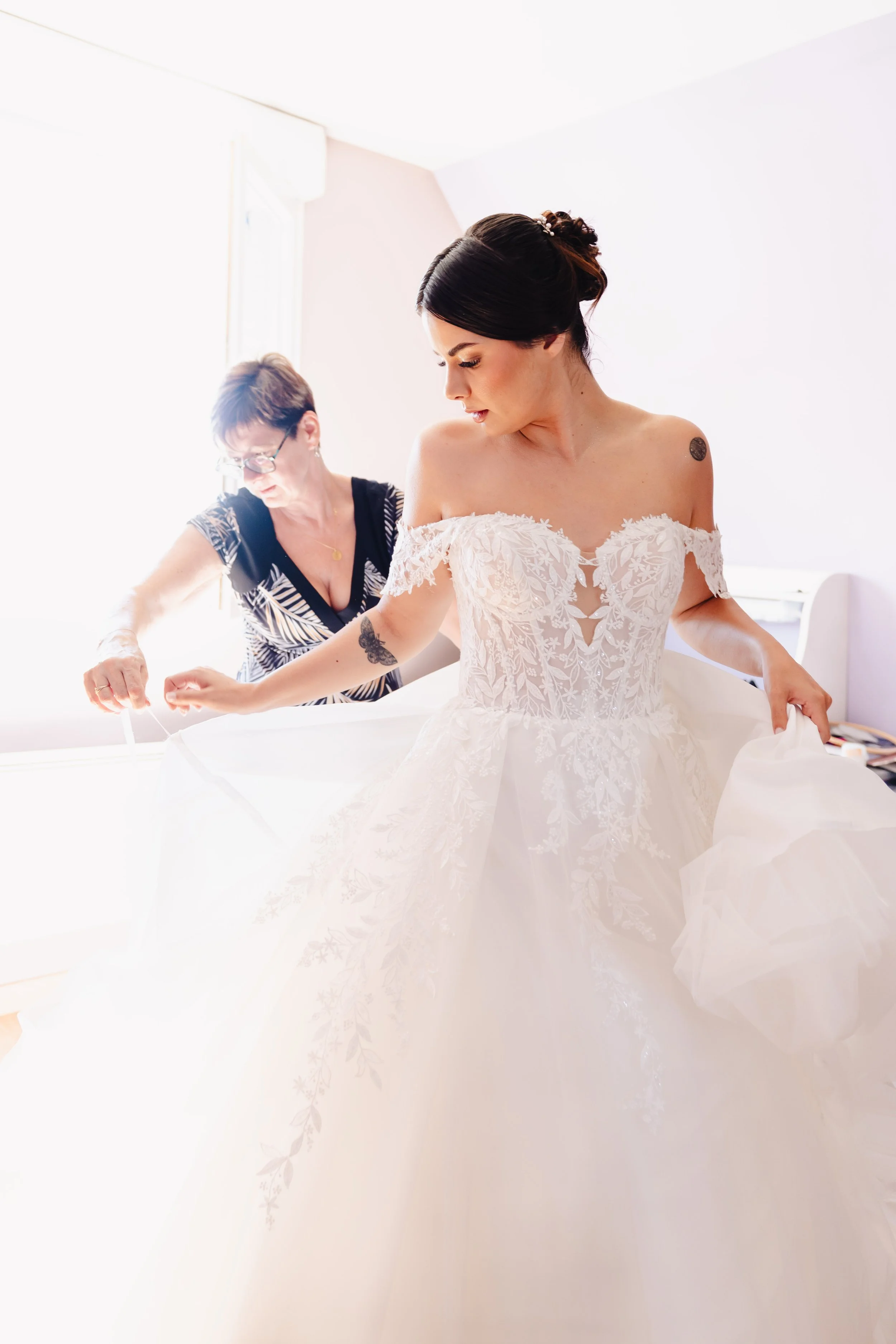 Une femme en robe de mariage se fait aider par une autre femme, probablement lors de la préparation pour un mariage. La femme en robe de mariage est en train de ajuster sa robe tandis que l'autre femme l'aide.