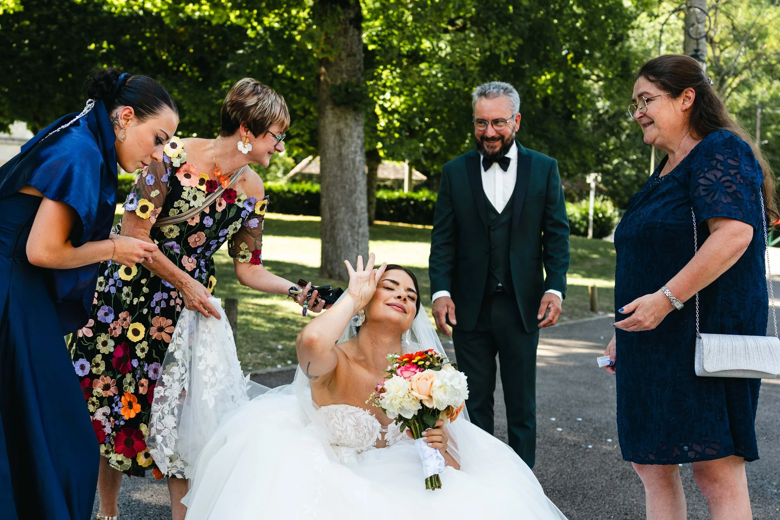 Une mariée en robe blanche souriante et faisant un signe de la main, entourée de quatre personnes dans un parc, lors d'un mariage.