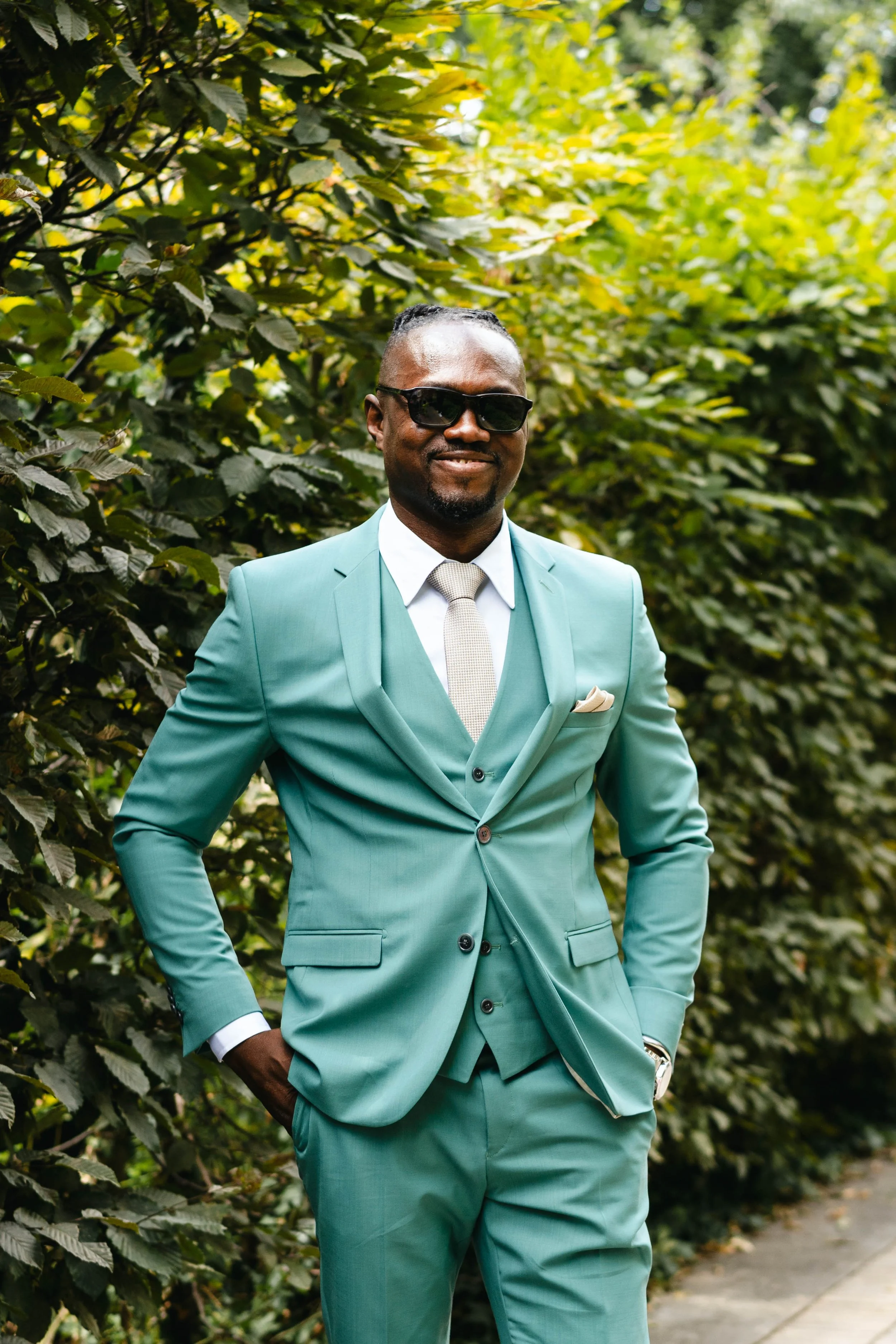 Homme en costume vert foncé avec une cravate beige, portant des lunettes de soleil, posant devant des arbres verts.