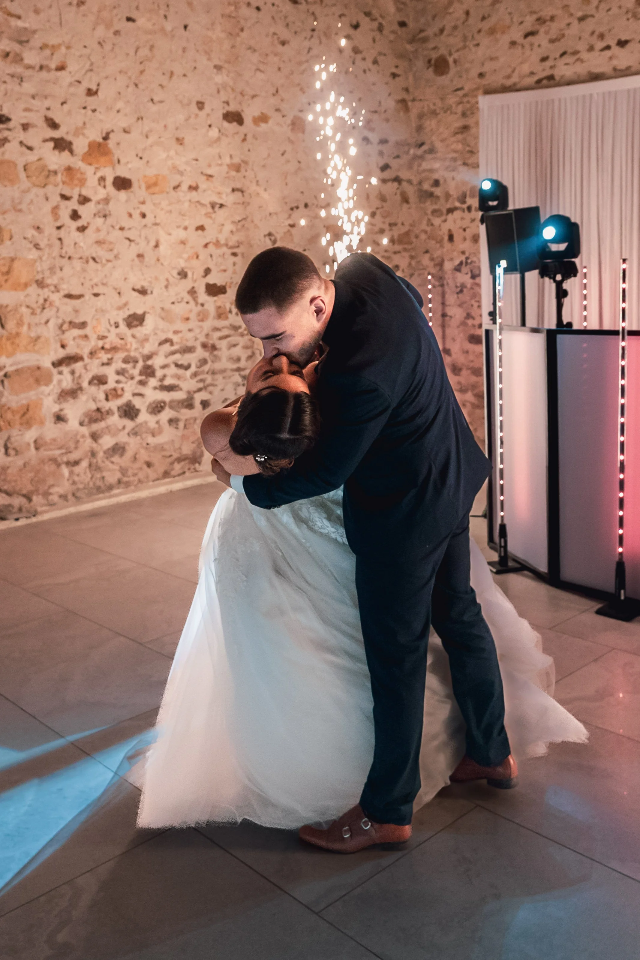 Un couple de mariés danse lors de leur mariage, dans une pièce avec mur en pierre et éclairage de fête.
