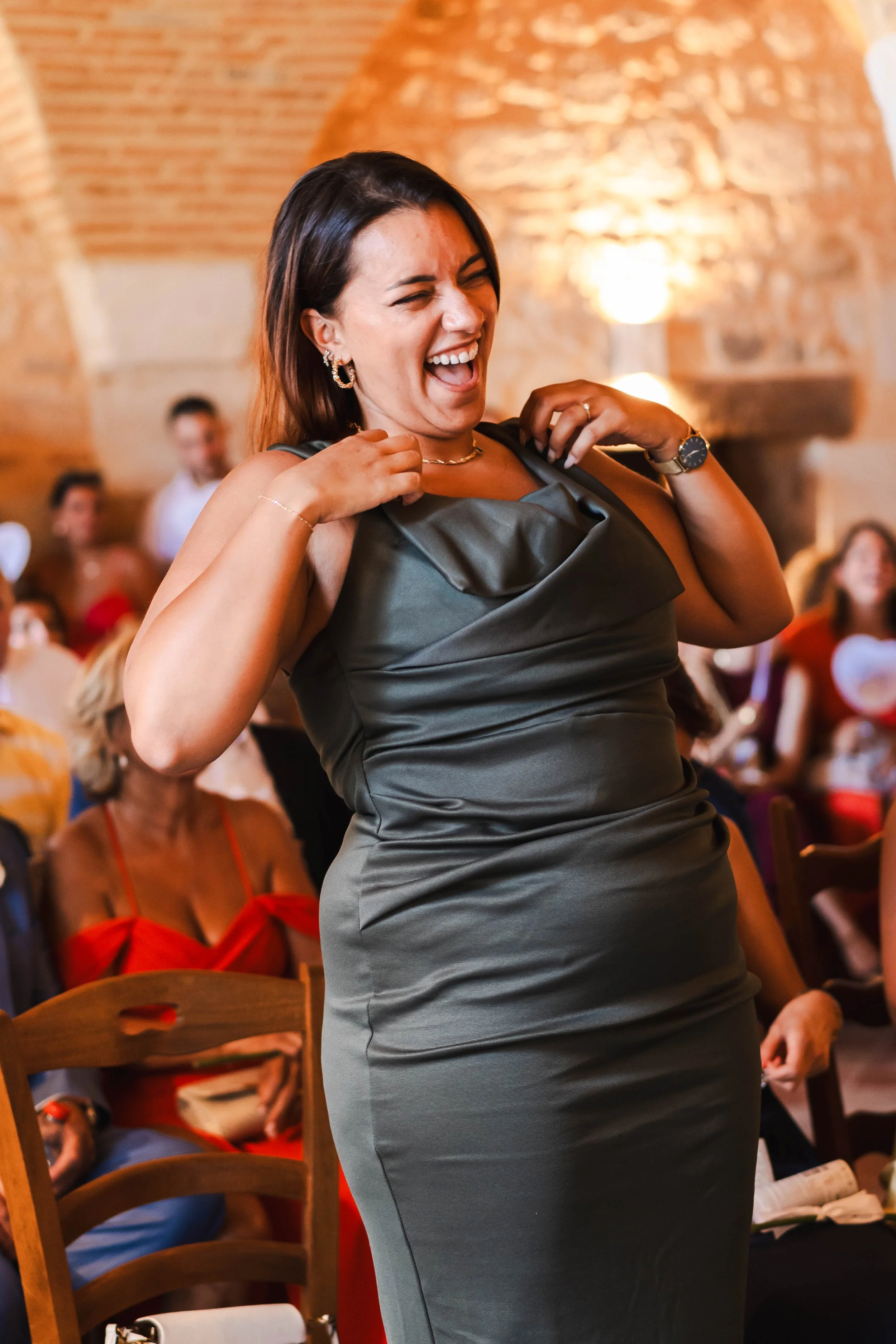 Femme souriante en robe grise, en train de danser lors d'un événement, avec des personnes assises en arrière-plan dans une salle avec une architecture en briques.