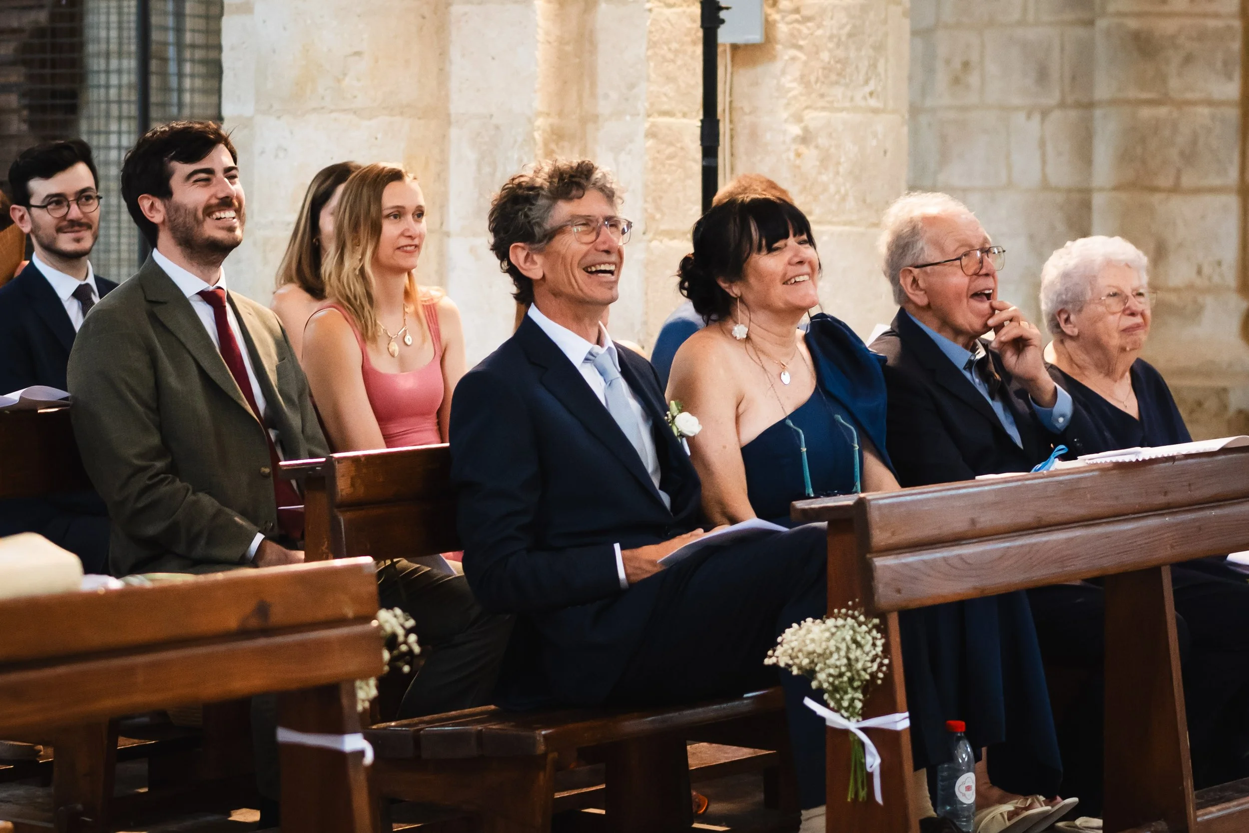 Groupe de personnes assises dans une église, souriantes et riant, probablement lors d'une cérémonie ou d'un mariage.
