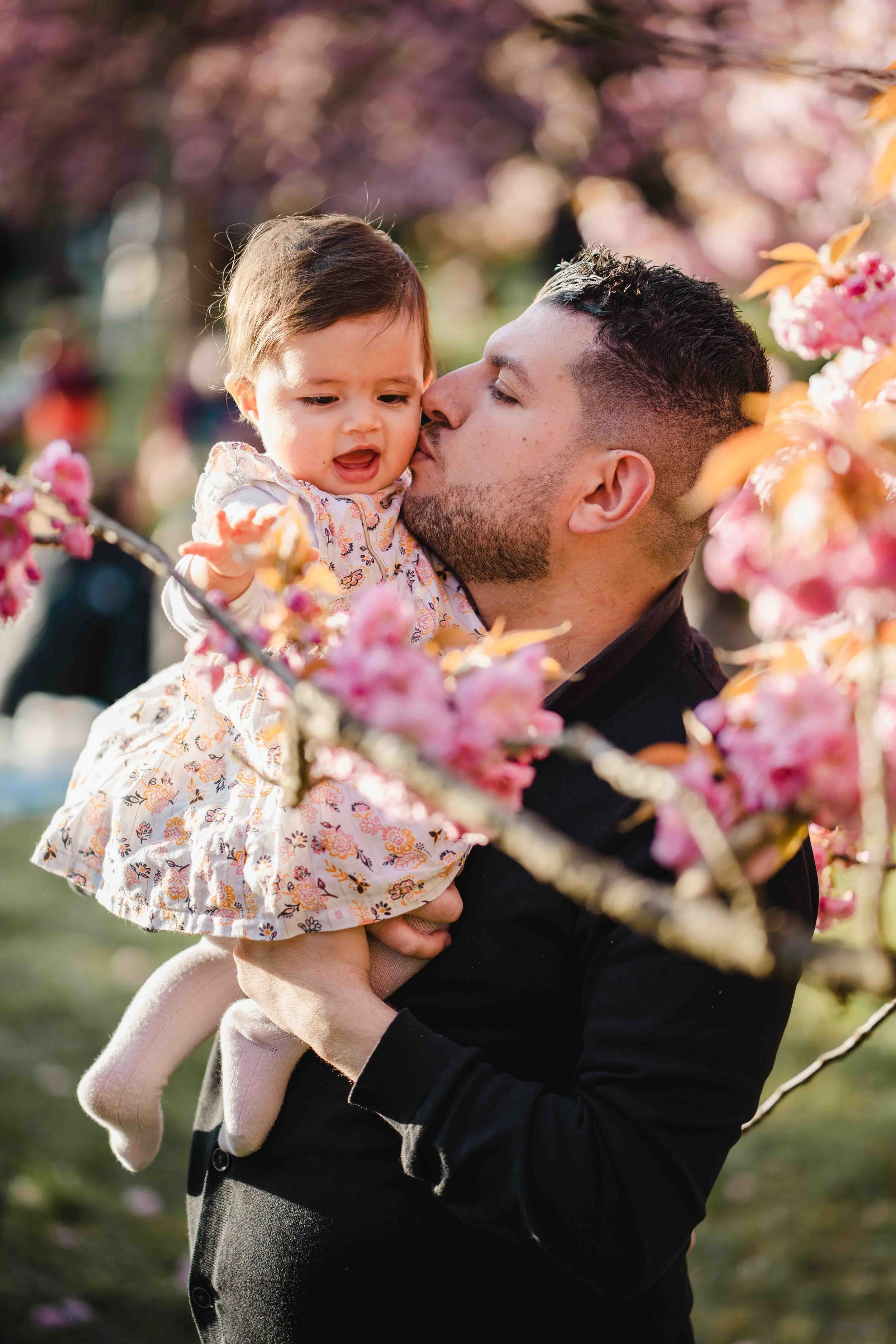 Un homme tient une petite fille dans ses bras, entourés de fleurs de cerisier en fleurs.