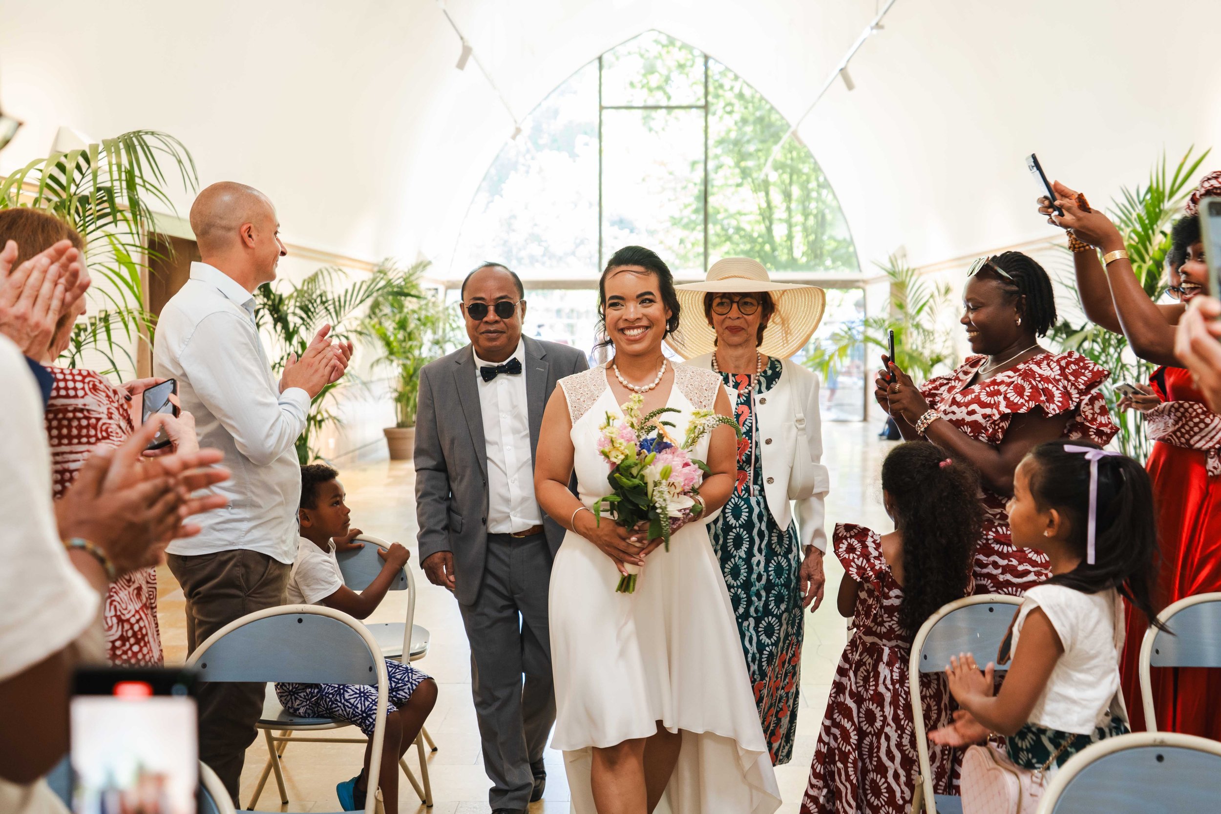 Une femme en robe blanche tenant un bouquet de fleurs sourit au centre d'une cérémonie. Autour, des personnes applaudent et prennent des photos.
