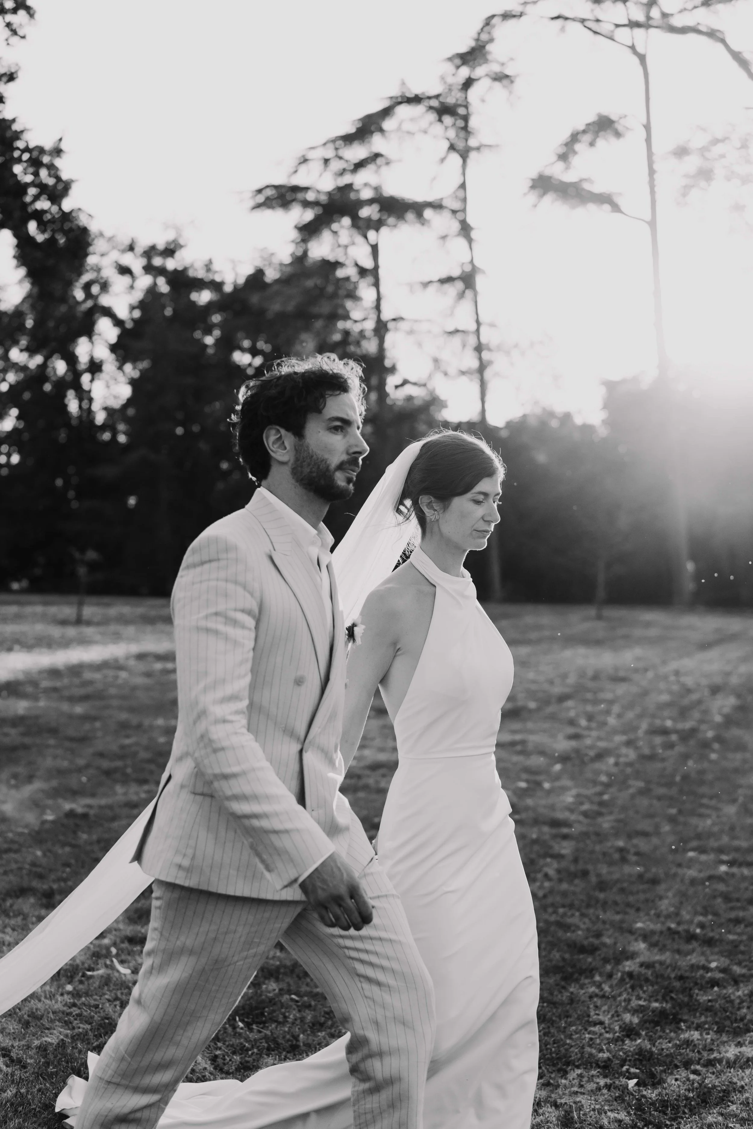 Un couple de mariés marche dans un parc en plein air, en noir et blanc, avec des arbres en arrière-plan, pendant le coucher du soleil.