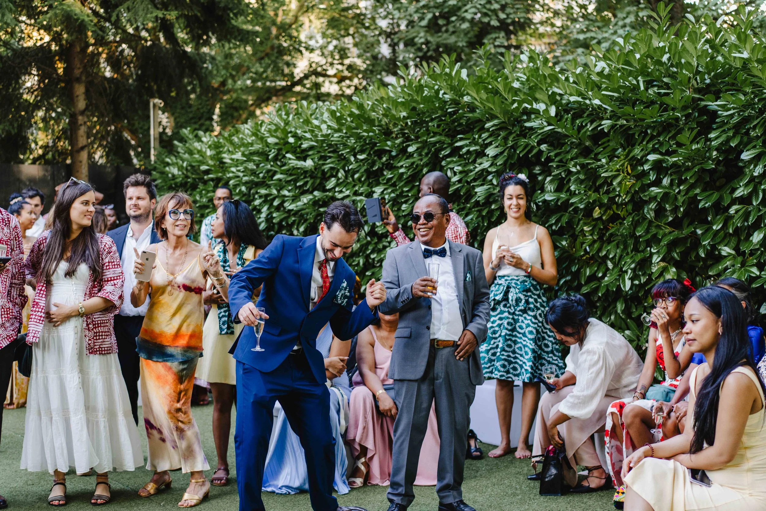 Groupe de personnes lors d'une fête en extérieur, certains dansent et d'autres regardent, avec un fond de haie verte et de grands arbres.