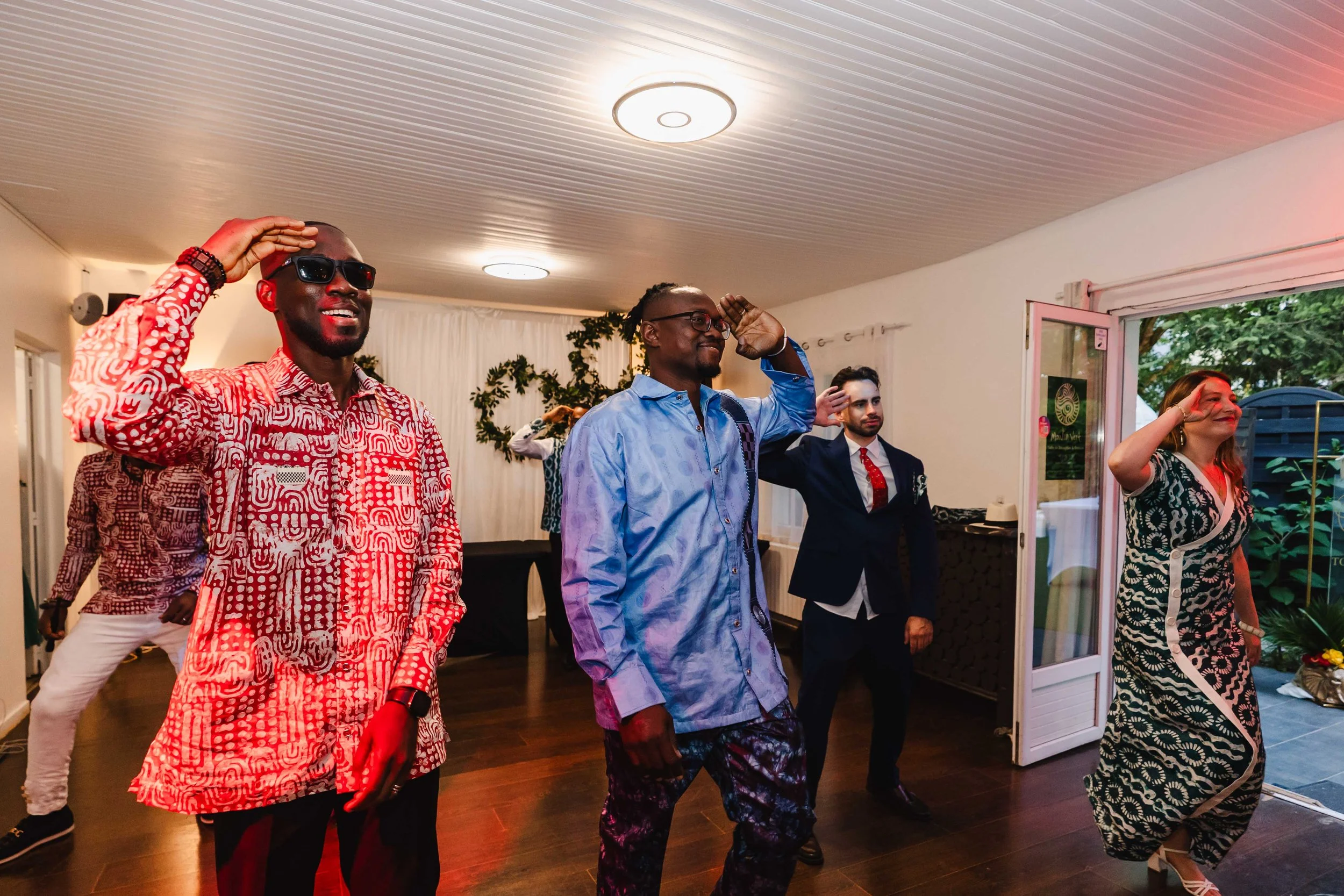 Groupe de personnes dansant lors d'une célébration dans une salle intérieure, avec une porte ouverte sur un espace extérieur vert.