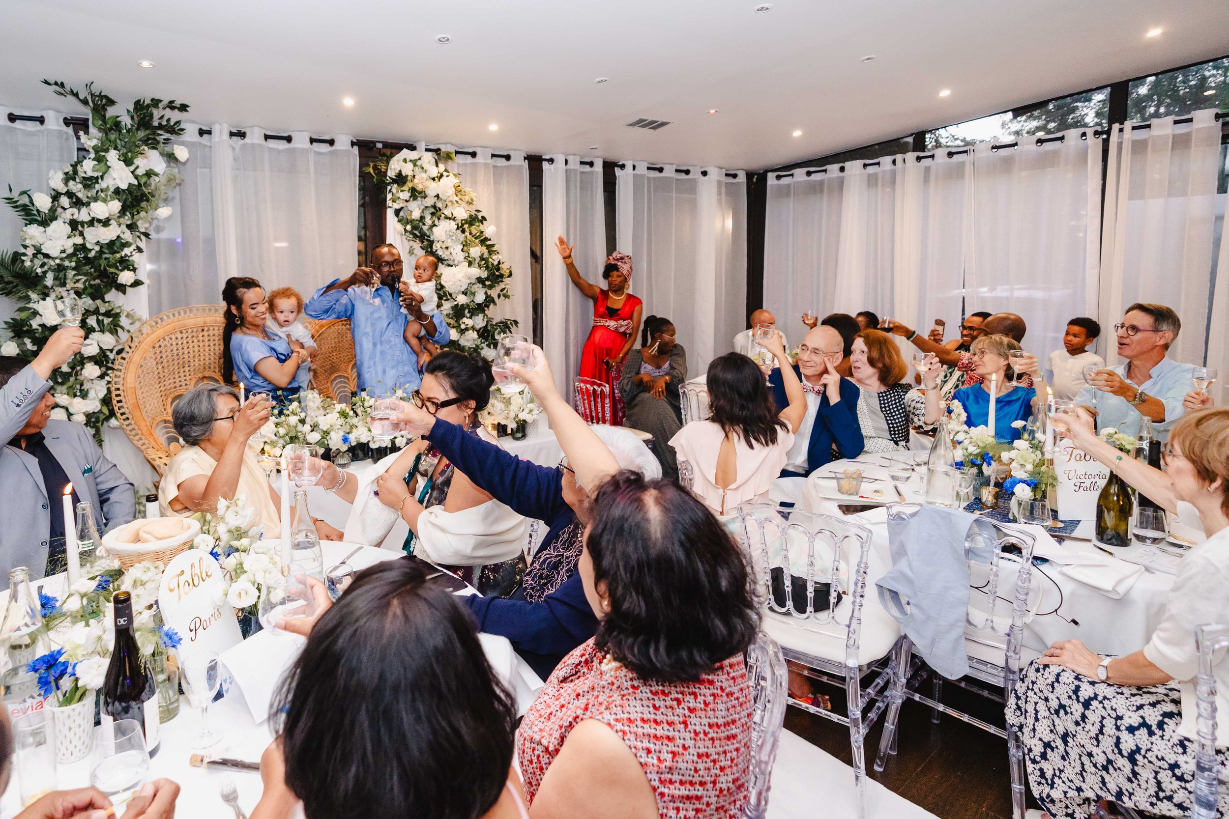 Un groupe de personnes assises autour d'une table lors d'une réception ou fête, célébrant avec des toasts. En arrière-plan, un ark décoratif avec des fleurs blanches et une femme en robe rouge dansant.
