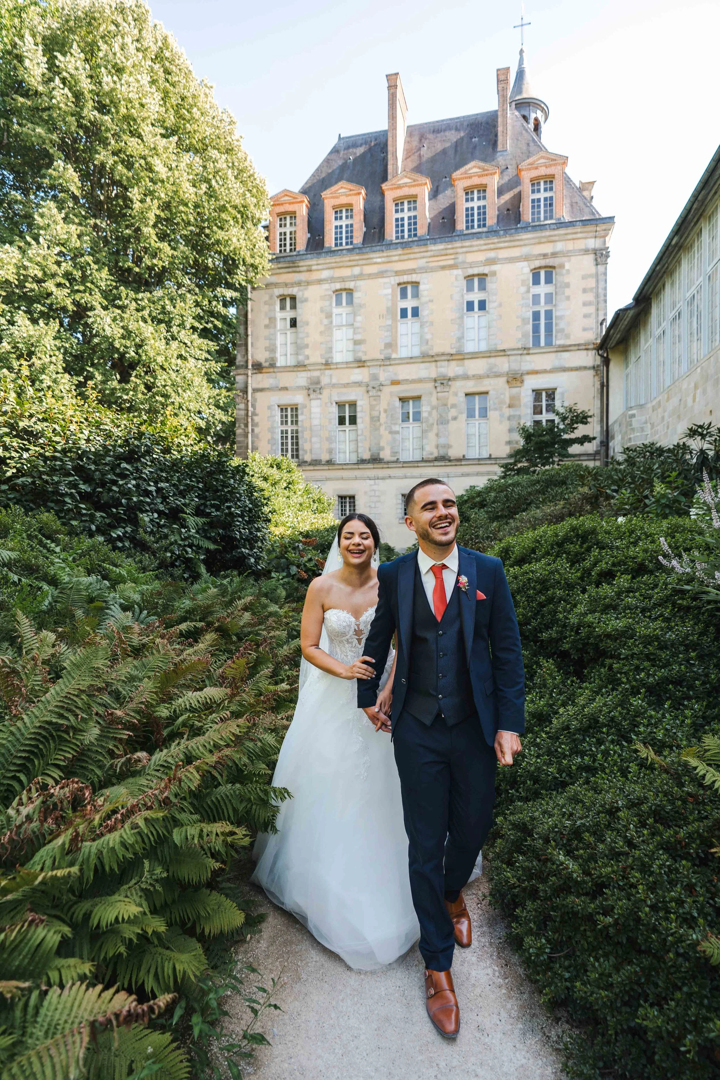 Un couple de mariés marche dans un jardin verdoyant avec un grand bâtiment ancien en arrière-plan, en plein jour. La mariée porte une robe blanche et le marié un costume bleu avec une cravate rouge.
