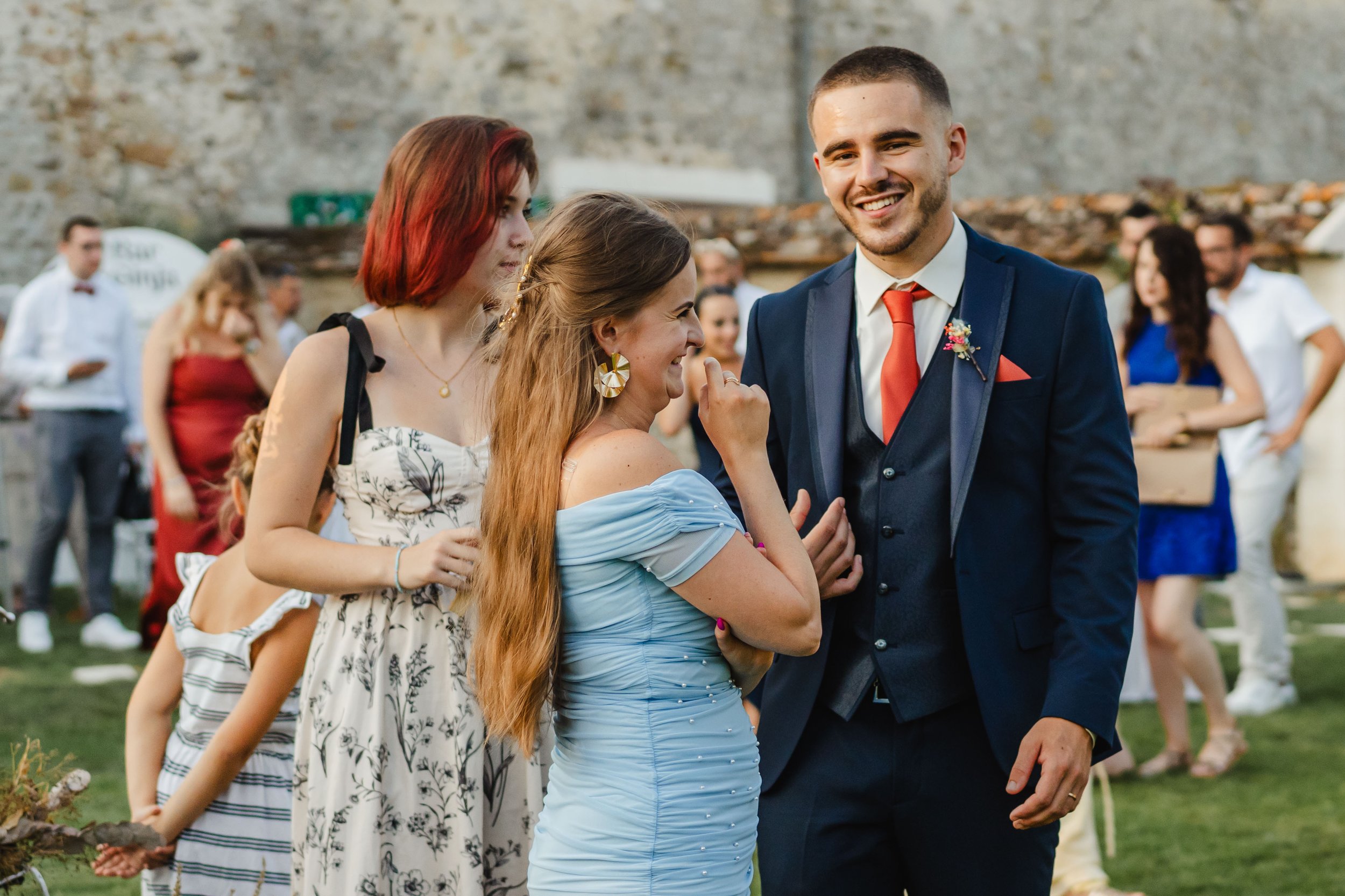 Groupe de personnes souriantes en tenue de soirée lors d'un événement en plein air, avec une structure en pierre en arrière-plan.