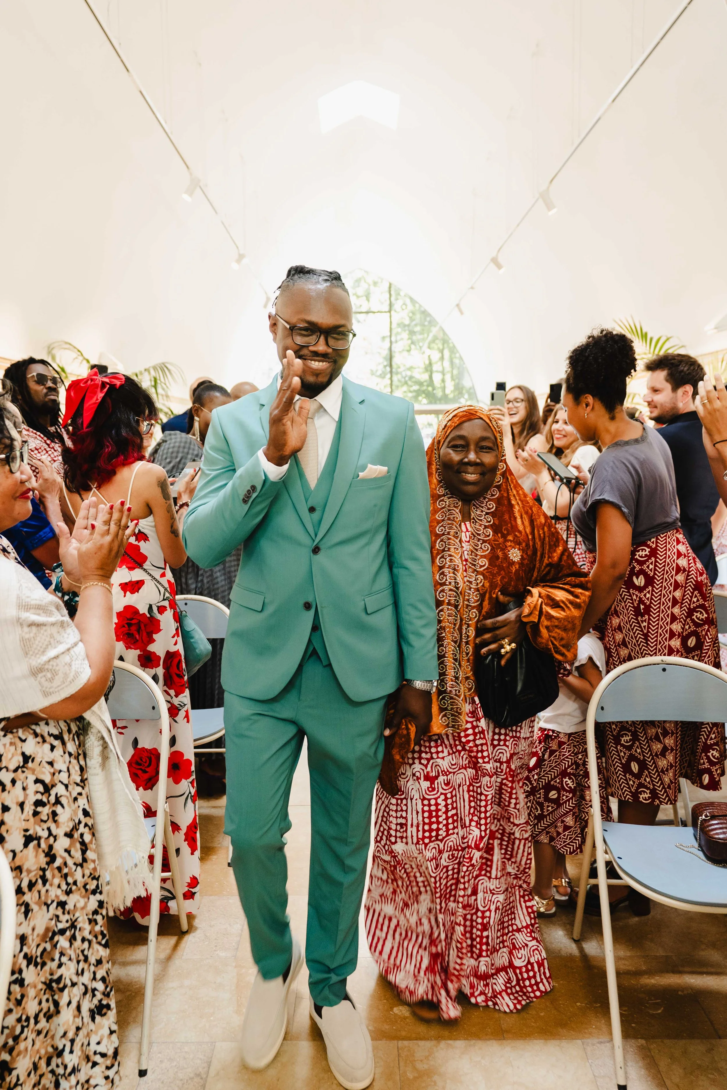 Un homme en costume turquoise et une femme en tenue traditionnelle debout au centre d'une salle, entourés de plusieurs personnes applaudissant, dans un lieu lumineux aux grandes fenêtres.