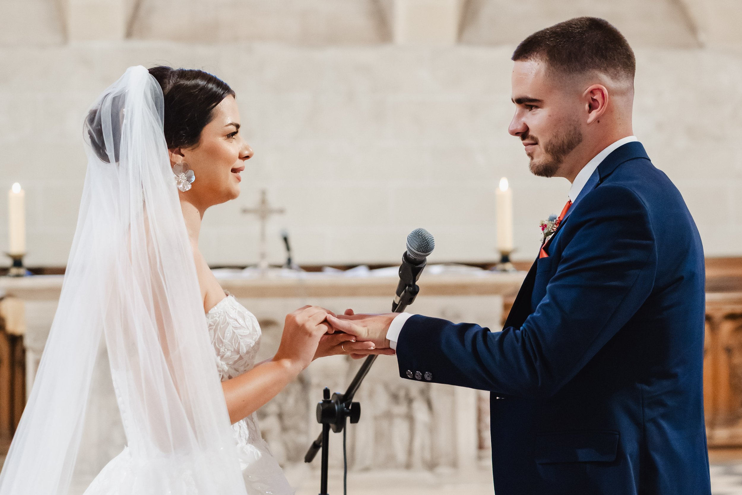 Un couple de mariés échangeant leurs vœux lors de leur mariage dans une église. La mariée porte une robe blanche avec un voile, et le marié porte un costume bleu. Ils se tiennent la main devant un pupitre, avec une croix et des bougies en arrière-pla