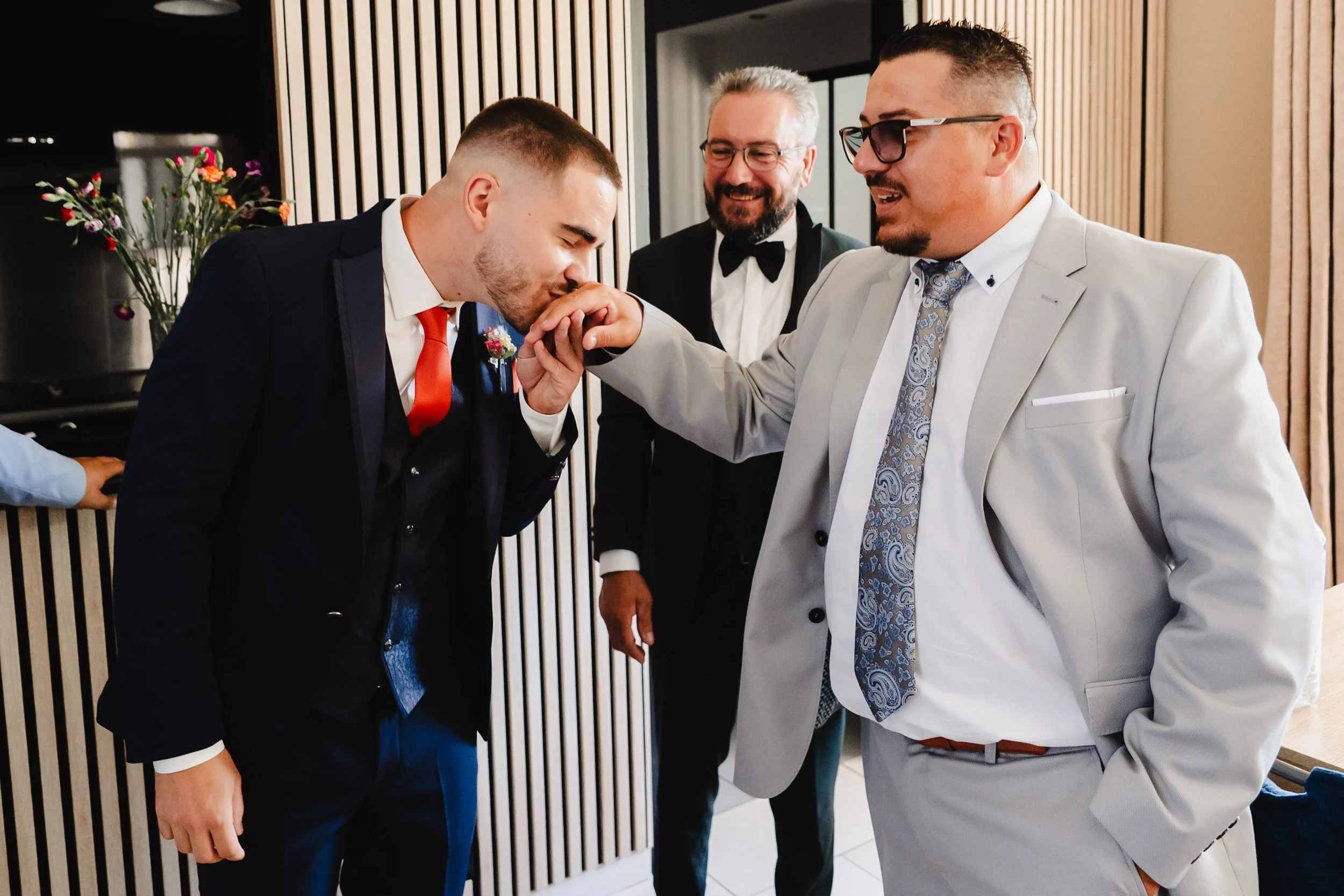 Trois hommes en costume lors d'une occasion spéciale, l'un d'eux embrasse la main de l'autre.