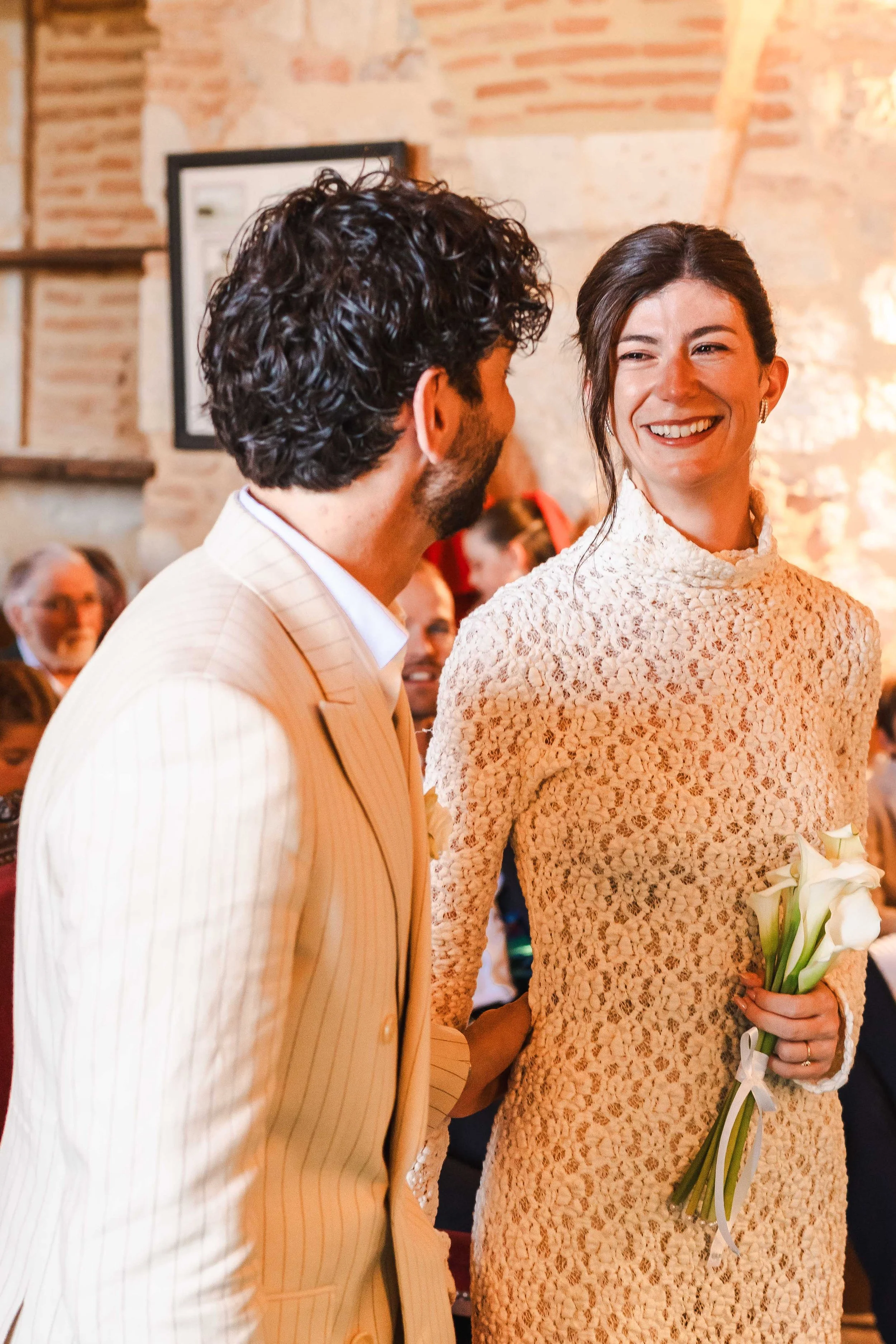 Un couple marié lors d'une cérémonie, la femme tenant un bouquet de fleurs avec une robe en dentelle et l'homme portant un costume à rayures, entourés d'invités dans la salle de mariage de la mairie de Moirax avec des murs en pierre.