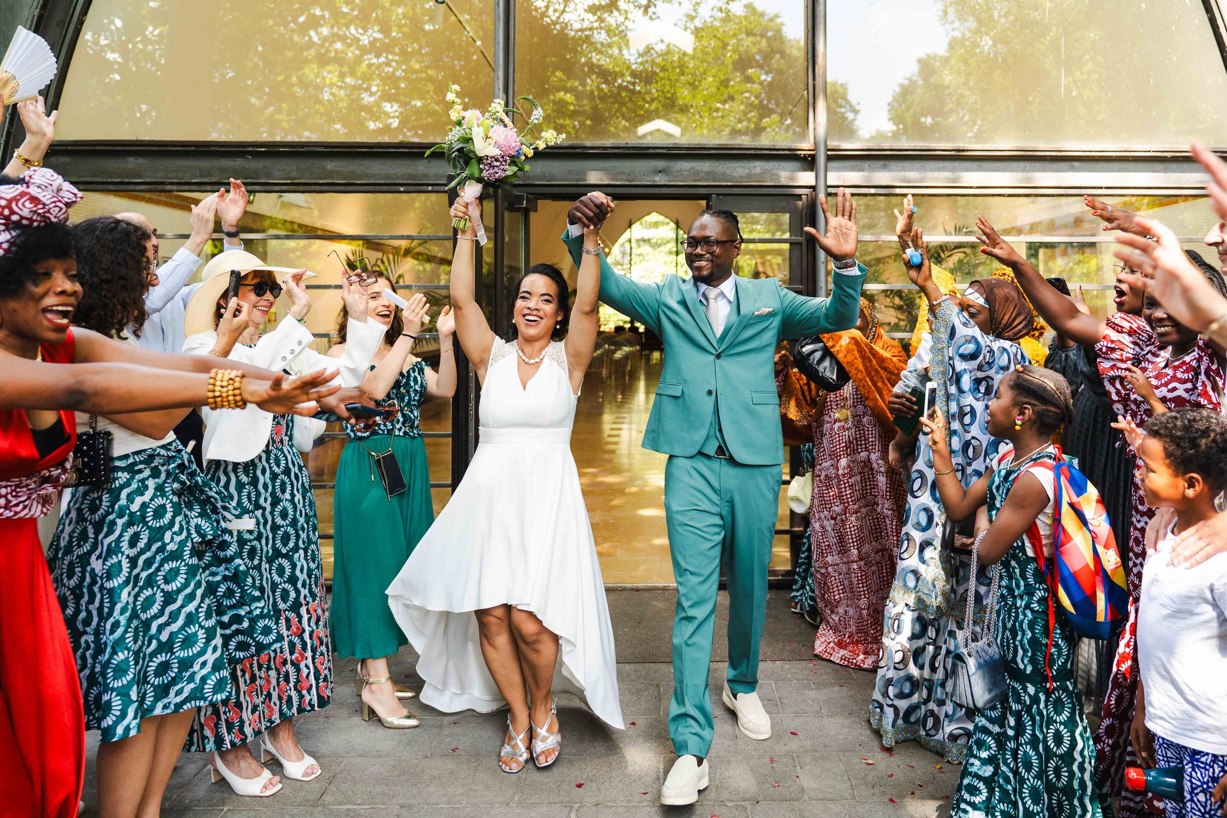 Un couple de mariage souriant, très heureuse, entrant dans un lieu de célébration, entouré d'invités qui l'acclament et prennent des photos. La femme porte une robe blanche, tandis que l'homme porte un costume bleu.
