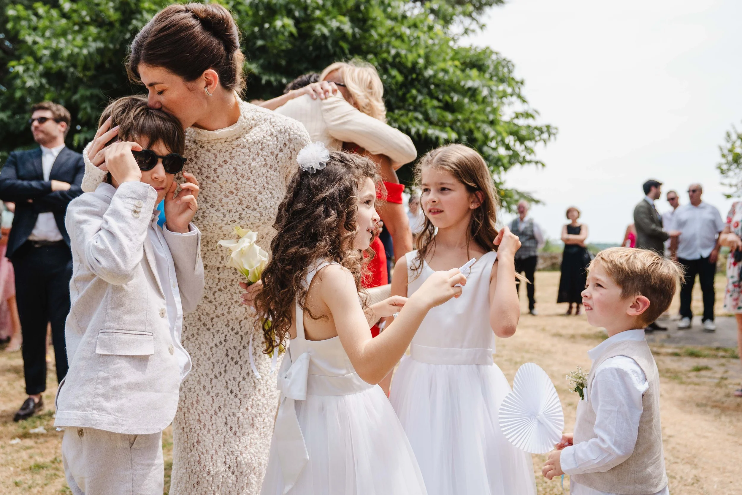Groupe d'enfants et d'adultes lors d'une fête en plein air, avec des enfants en vêtements blancs et des adultes en tenues formelles, sous un arbre.