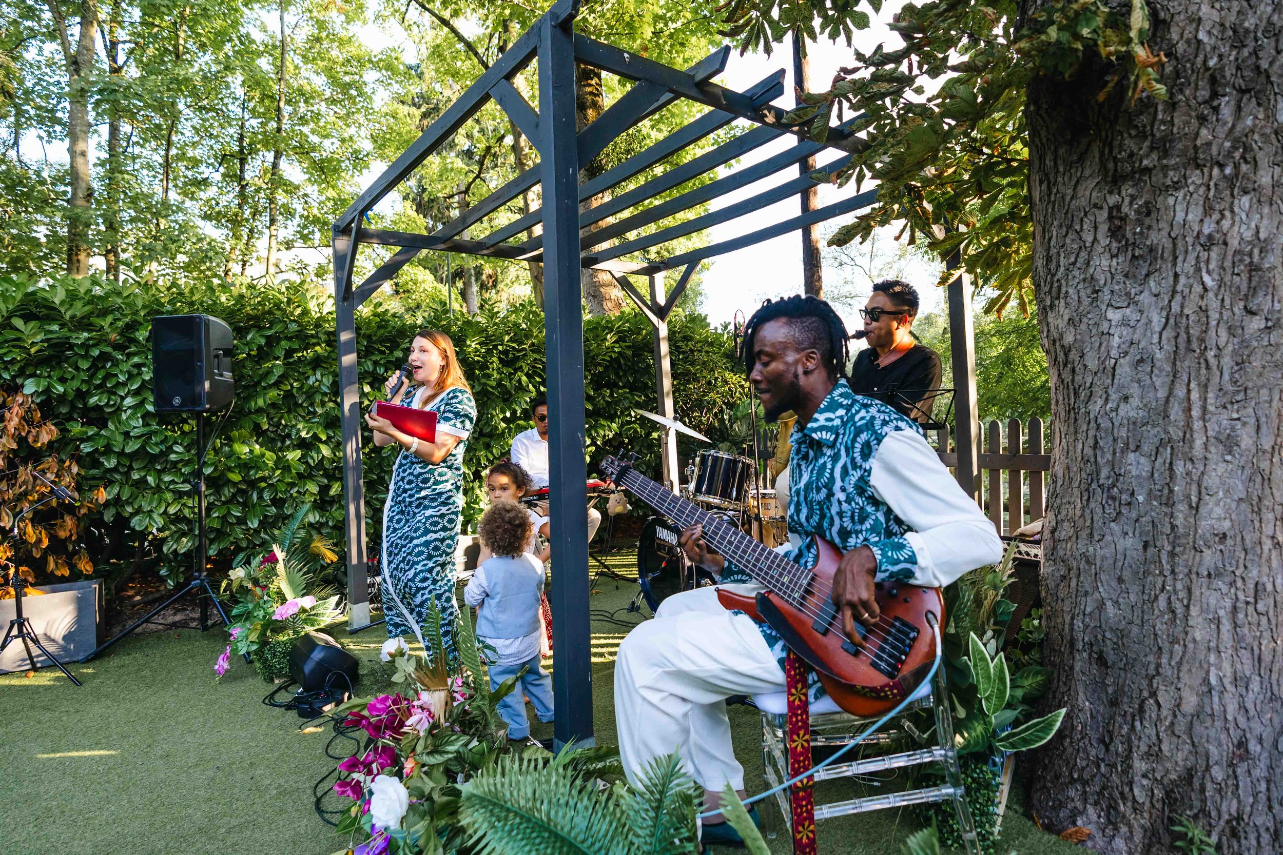 Une scène de concert en plein air avec plusieurs musiciens et un public. Un groupe de musiciens joue, dont un homme avec une guitare électrique et d'autres avec des instruments de percussion. Une femme chante avec un micro, accompagnée par deux enfan