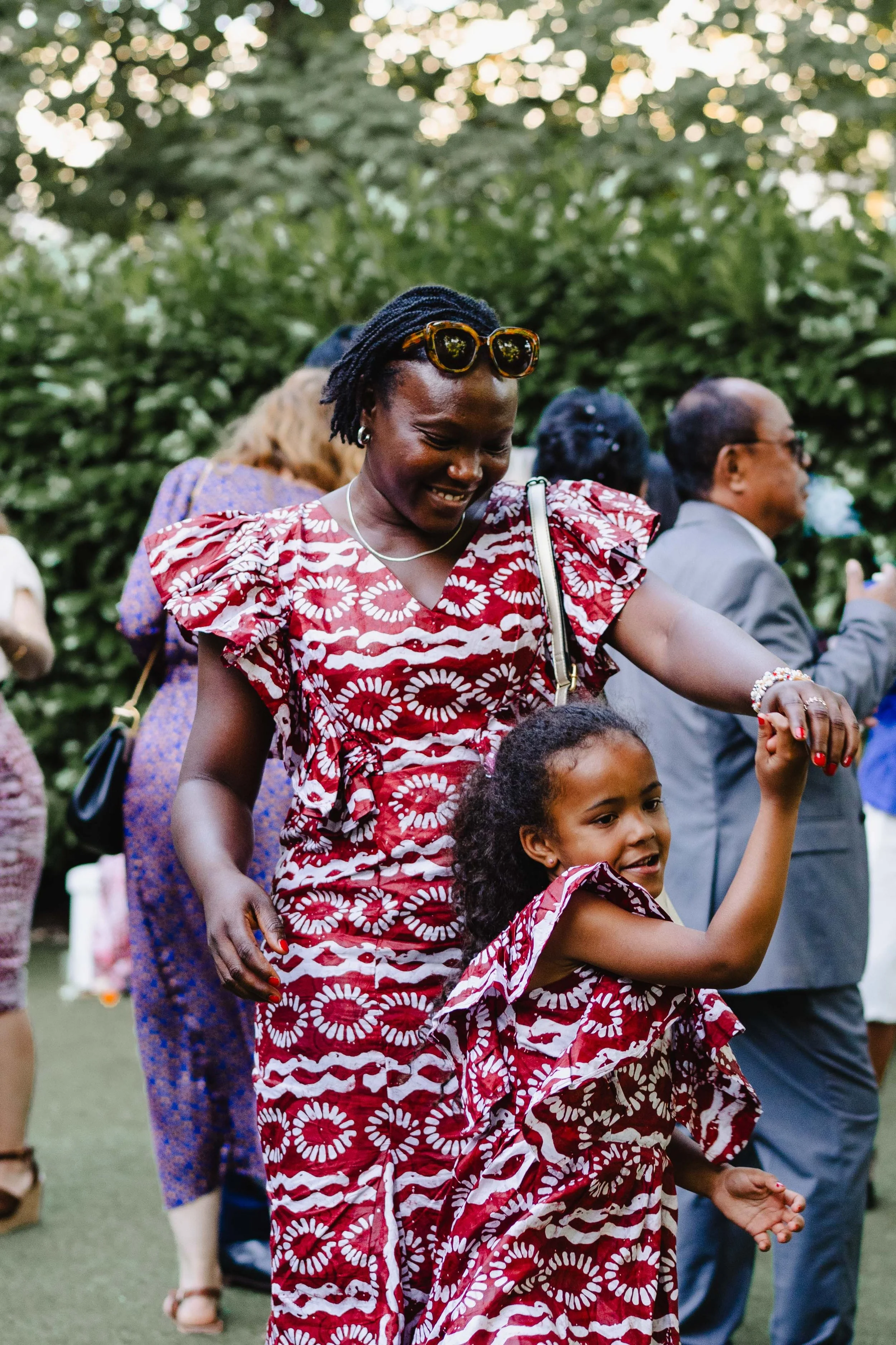 Une femme et une jeune fille dansent en plein air, entourées d'autres personnes. La femme porte une robe rouge à motifs blancs avec des sacs à main, et la jeune fille porte une robe coordonnée. Ils semblent heureux et profiter d'une occasion festive.
