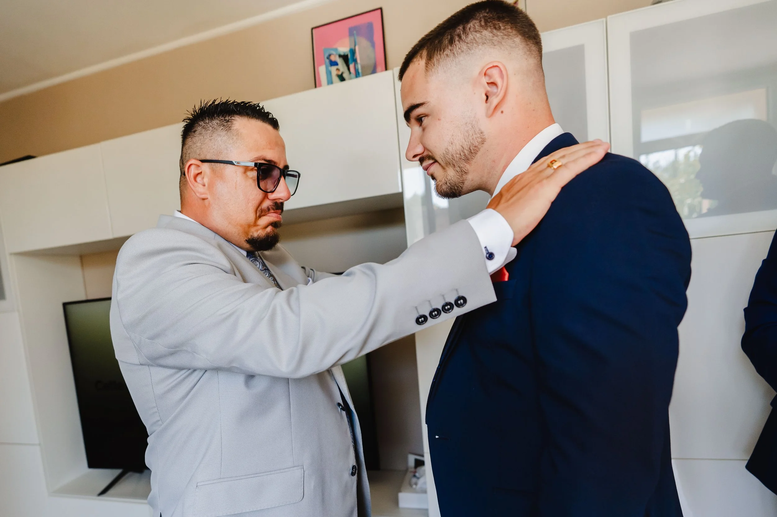 Deux hommes en costume se tiennent face à face dans une pièce, l'un touche l'autre sur l'épaule. L'homme à gauche porte des lunettes et un costume gris, l'homme à droite porte un costume bleu avec une chemise blanche et une cravate rouge.