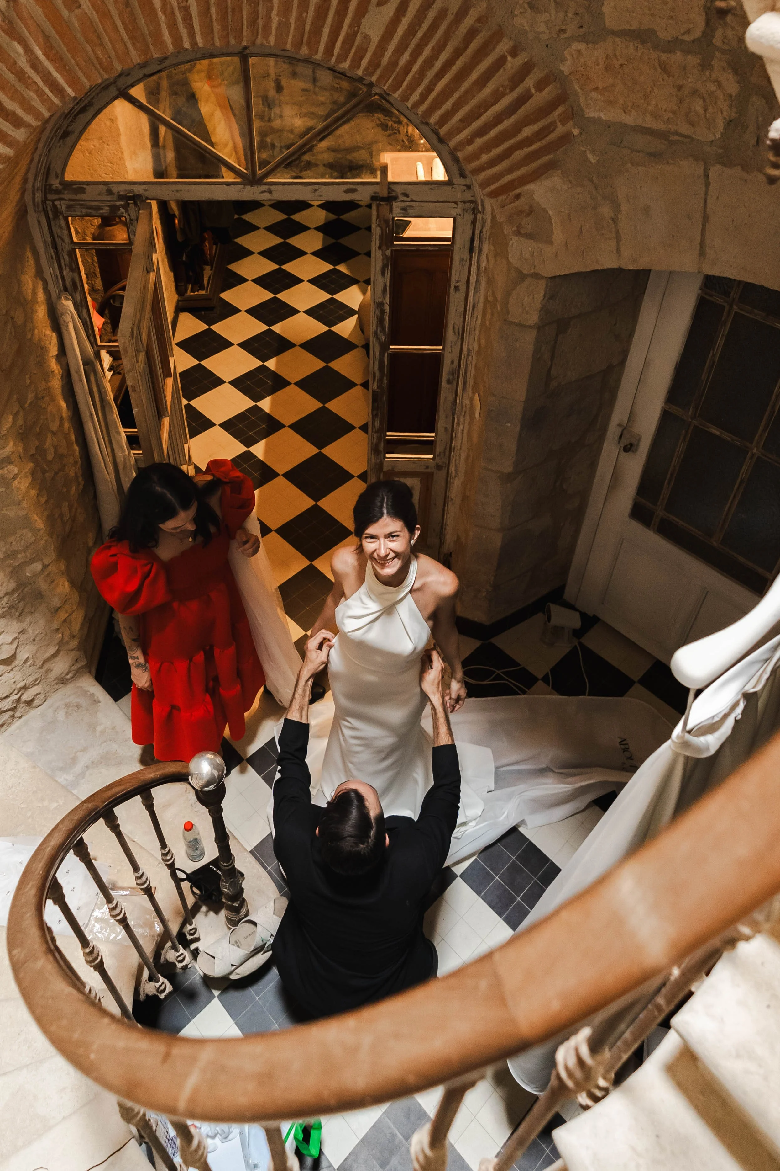 Une femme en robe blanche souriante lors d'une cérémonie, entourée de deux autres personnes, dans un intérieur aux murs en pierre et sol à carreaux noirs et blancs.